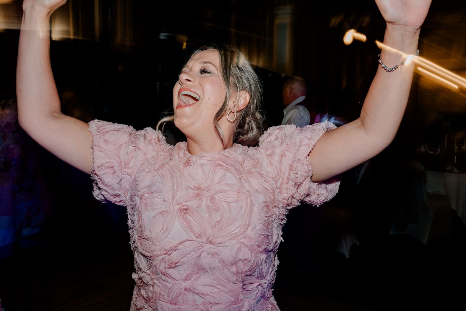 Woman in pink floral dress dancing joyfully with arms raised at a wedding reception.
