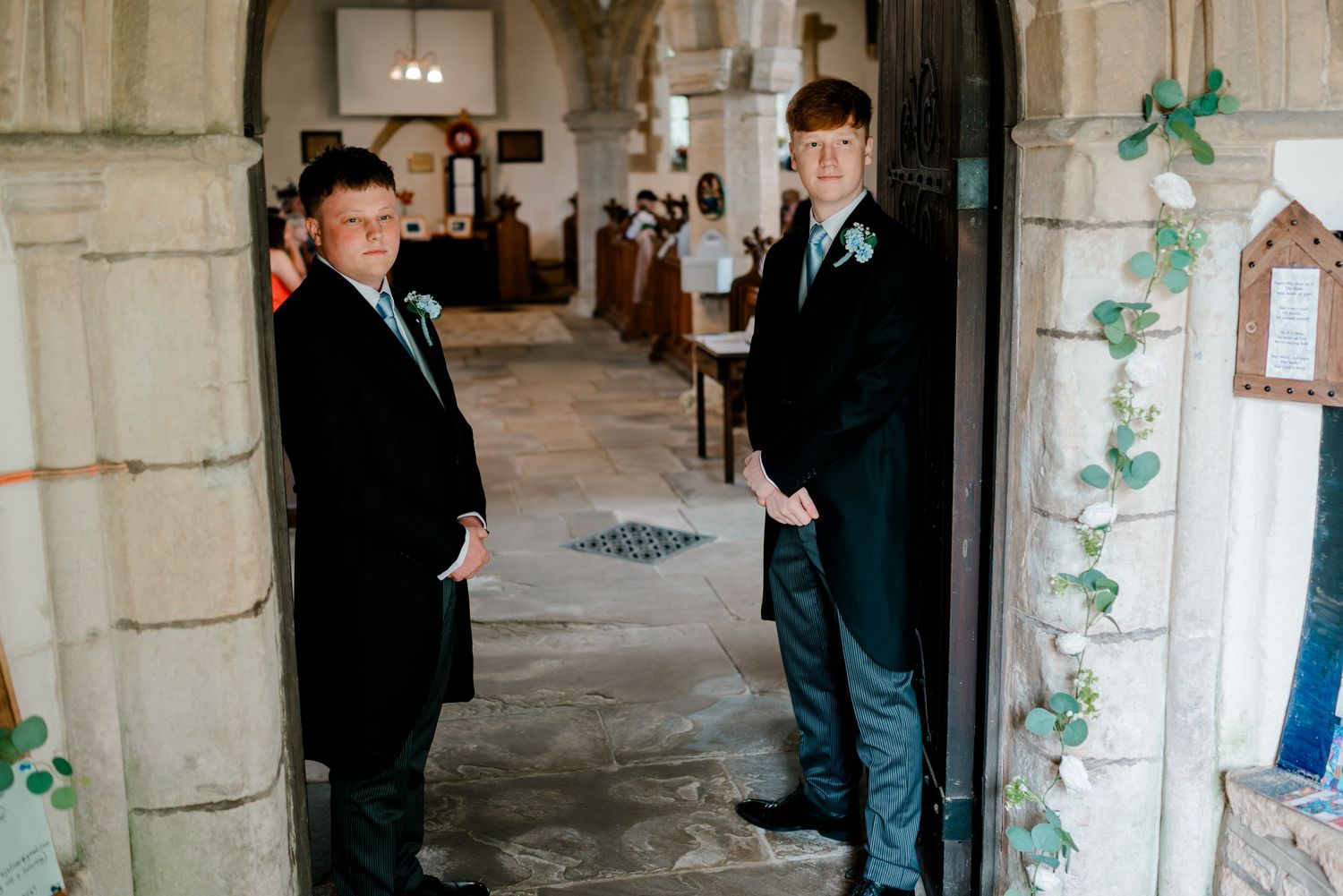 Two young men in suits stand at the entrance of a historic stone church, wearing boutonnieres.