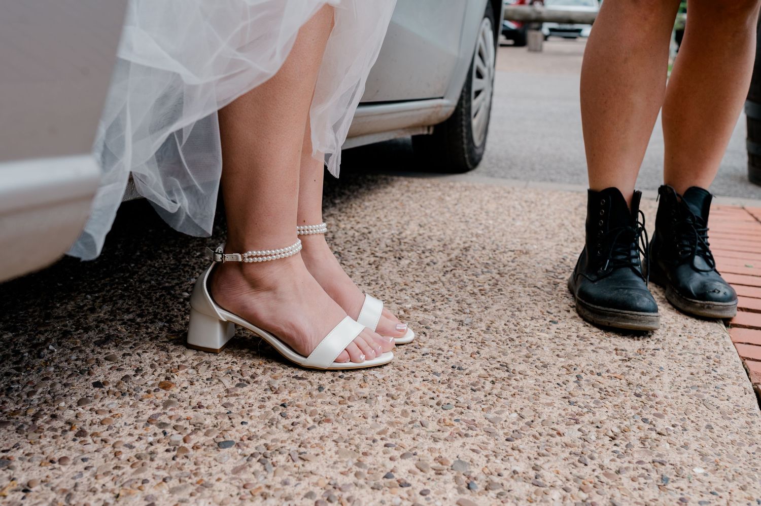 Close-up of bride's white strappy heels and groom's black boots on a paved surface near a car.