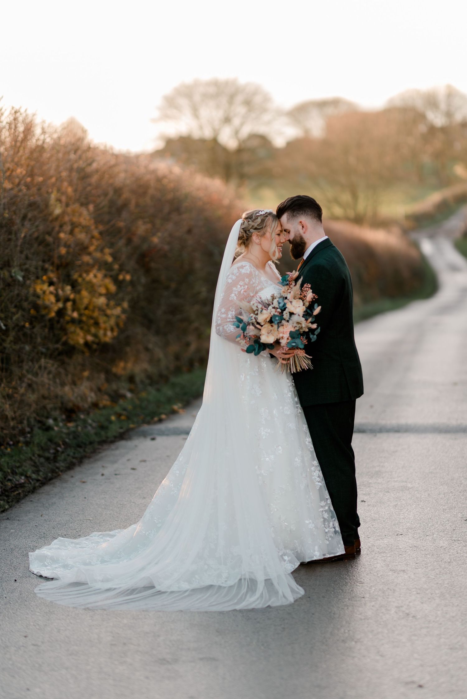 Bride and groom sharing romantic forehead touch on country lane at golden hour, bride holding pastel bouquet.