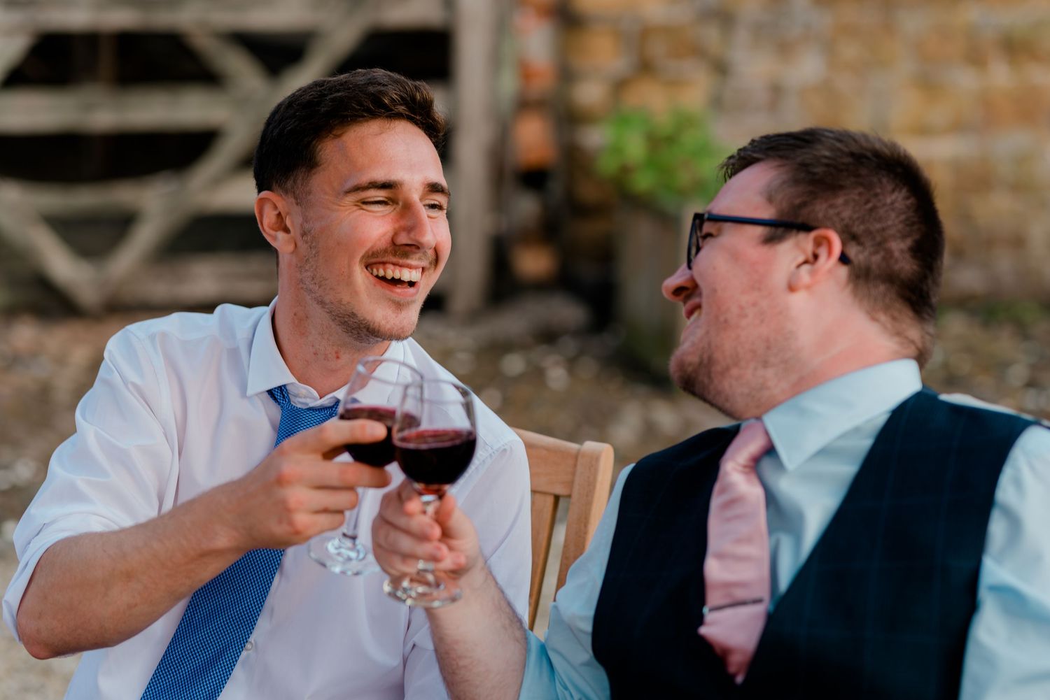 Two men laughing and toasting with red wine glasses at an outdoor wedding reception.