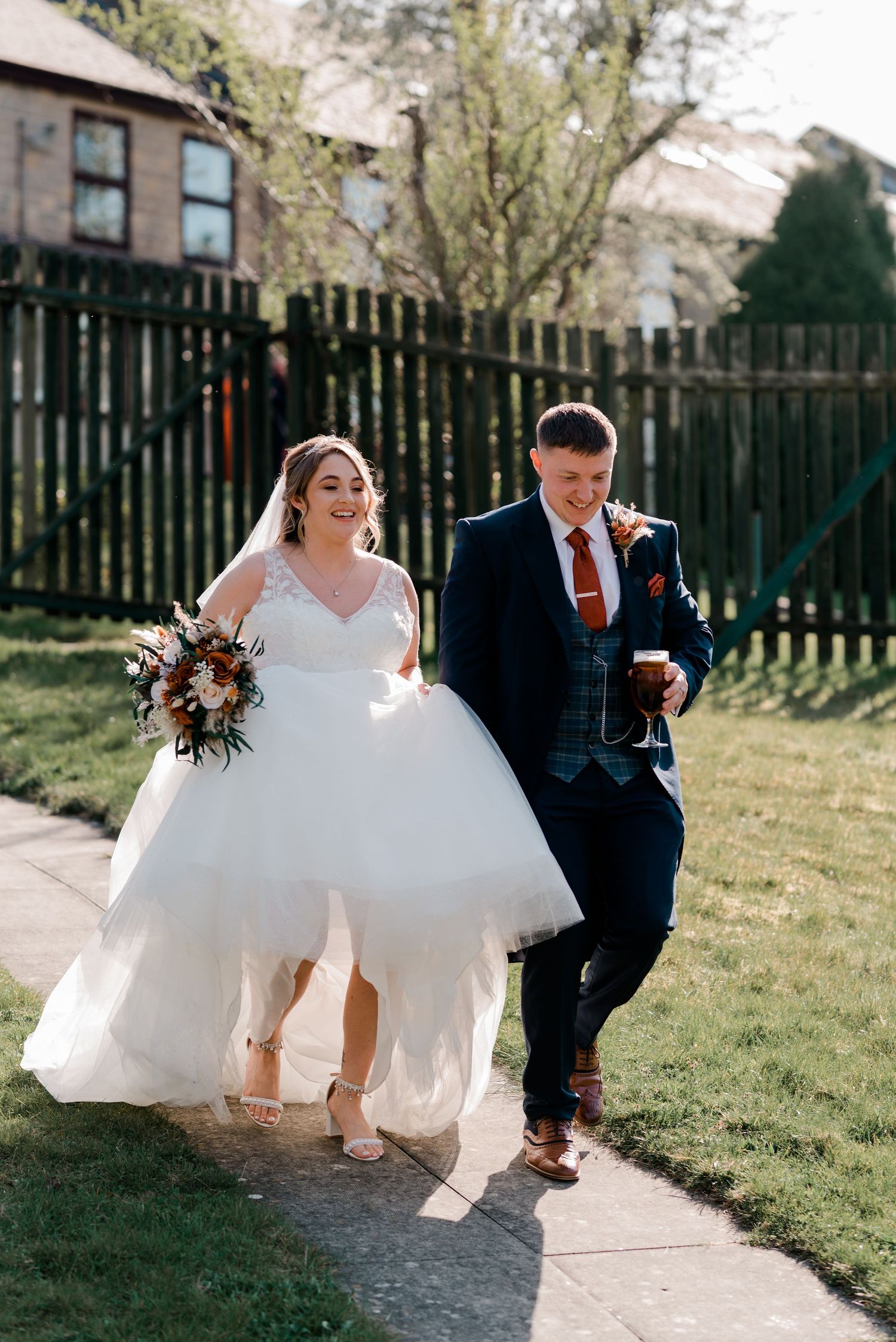 Happy bride in tulle gown and groom in navy suit walk along sunny path beside wooden fence.