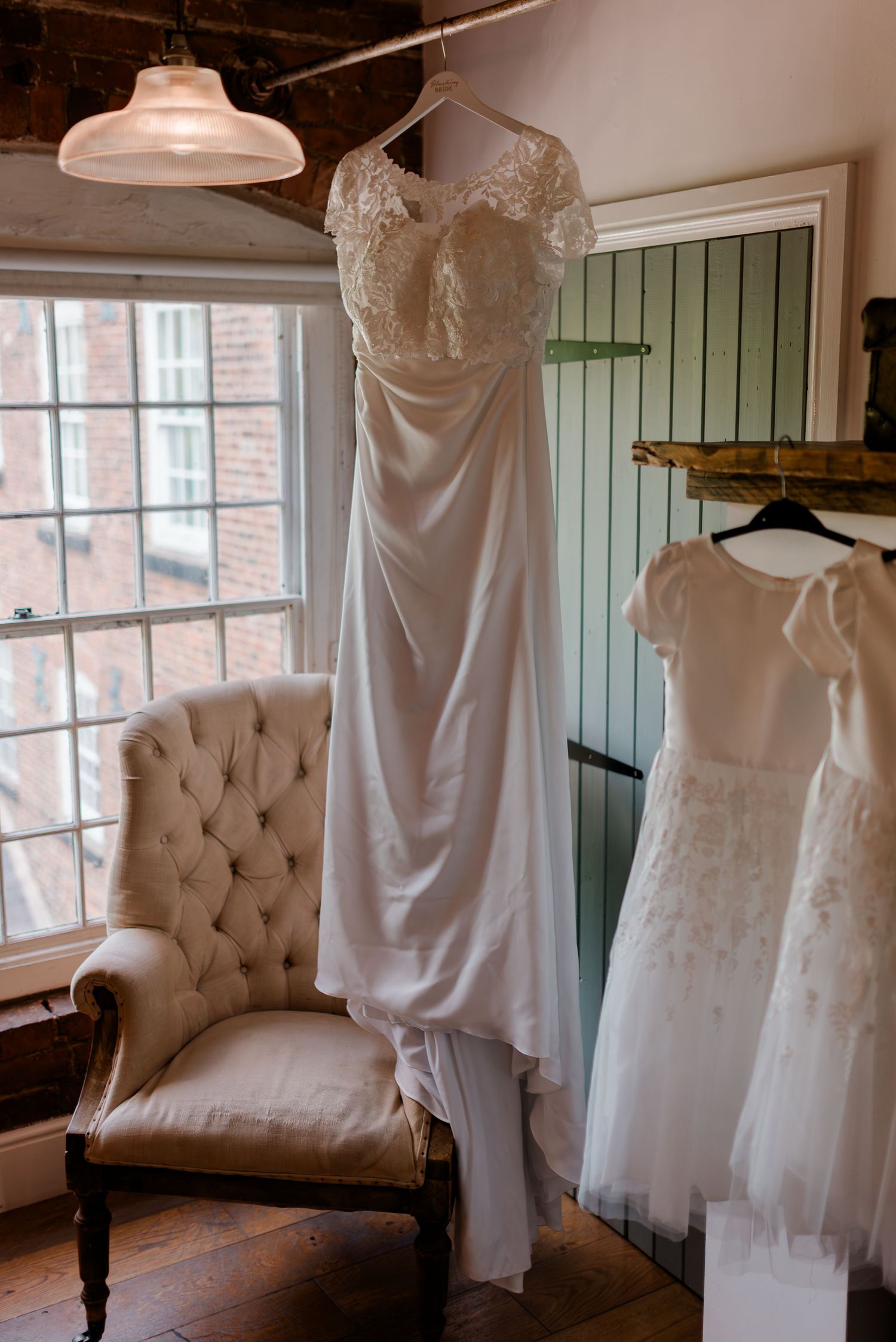 Delicate white wedding dresses and gowns displayed on hangers next to an elegant tufted armchair in a rustic bridal suite.