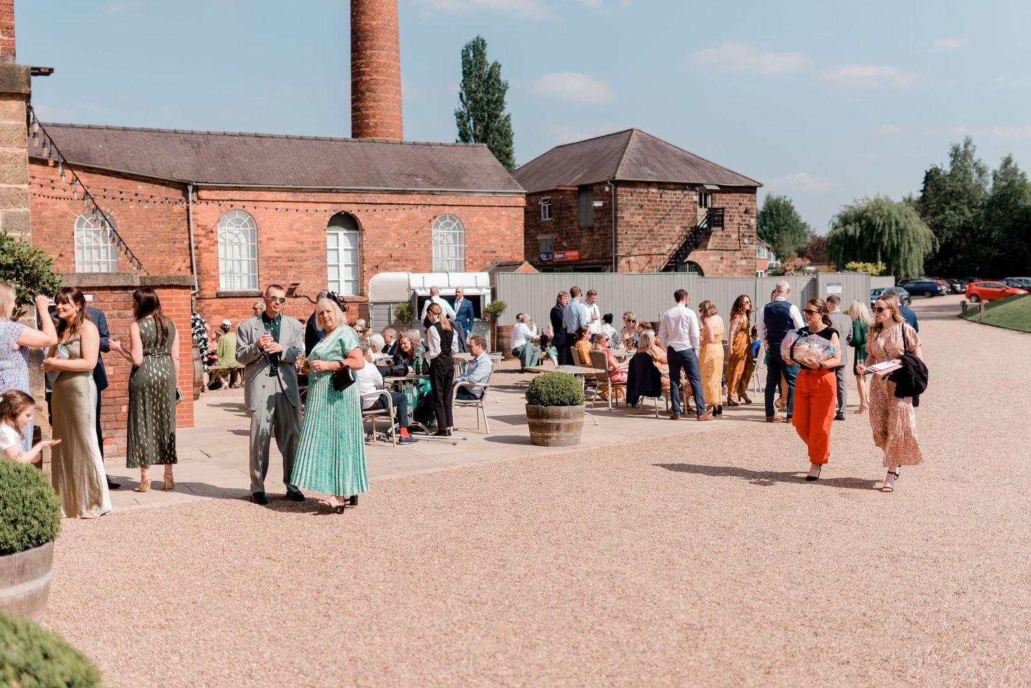 Panoramic view of wedding guests gathered in sunny courtyard of historic brick building complex.