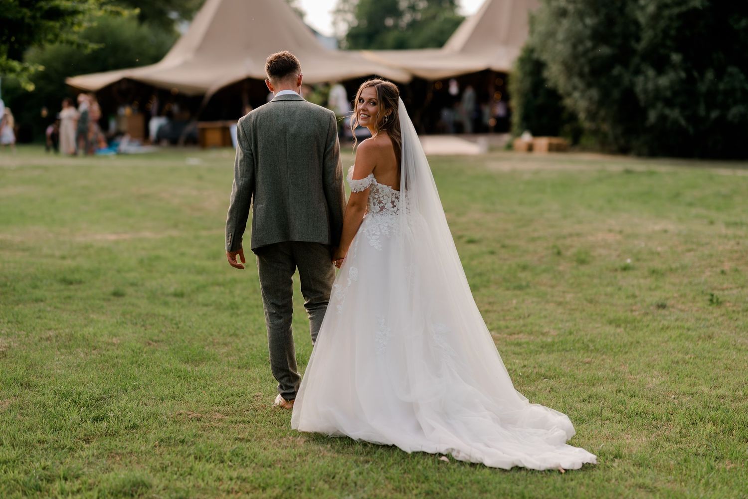 Bride and groom walking hand in hand across green lawn toward tipi wedding reception venue.