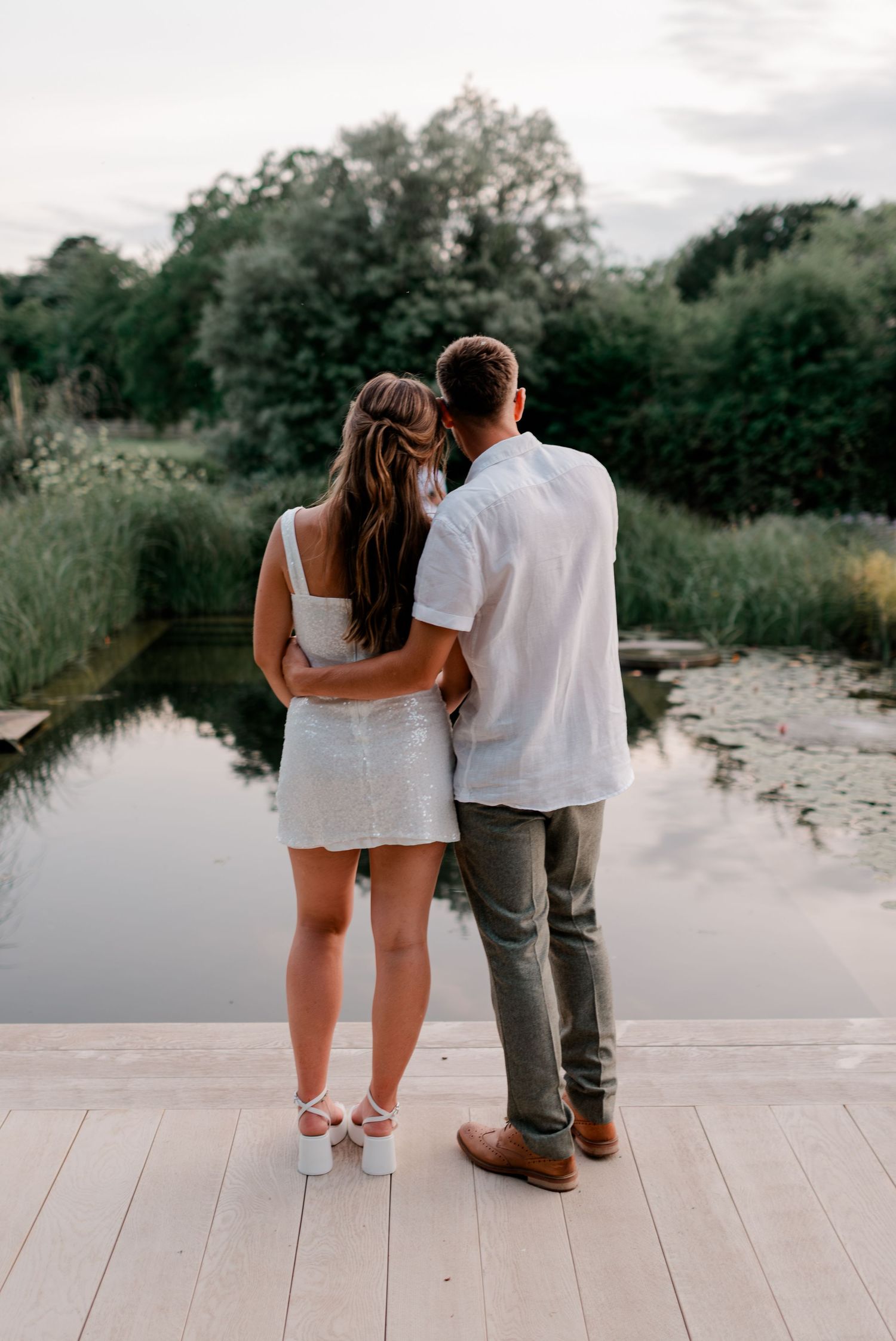 A couple sits together on a wooden deck overlooking a serene pond at sunset.