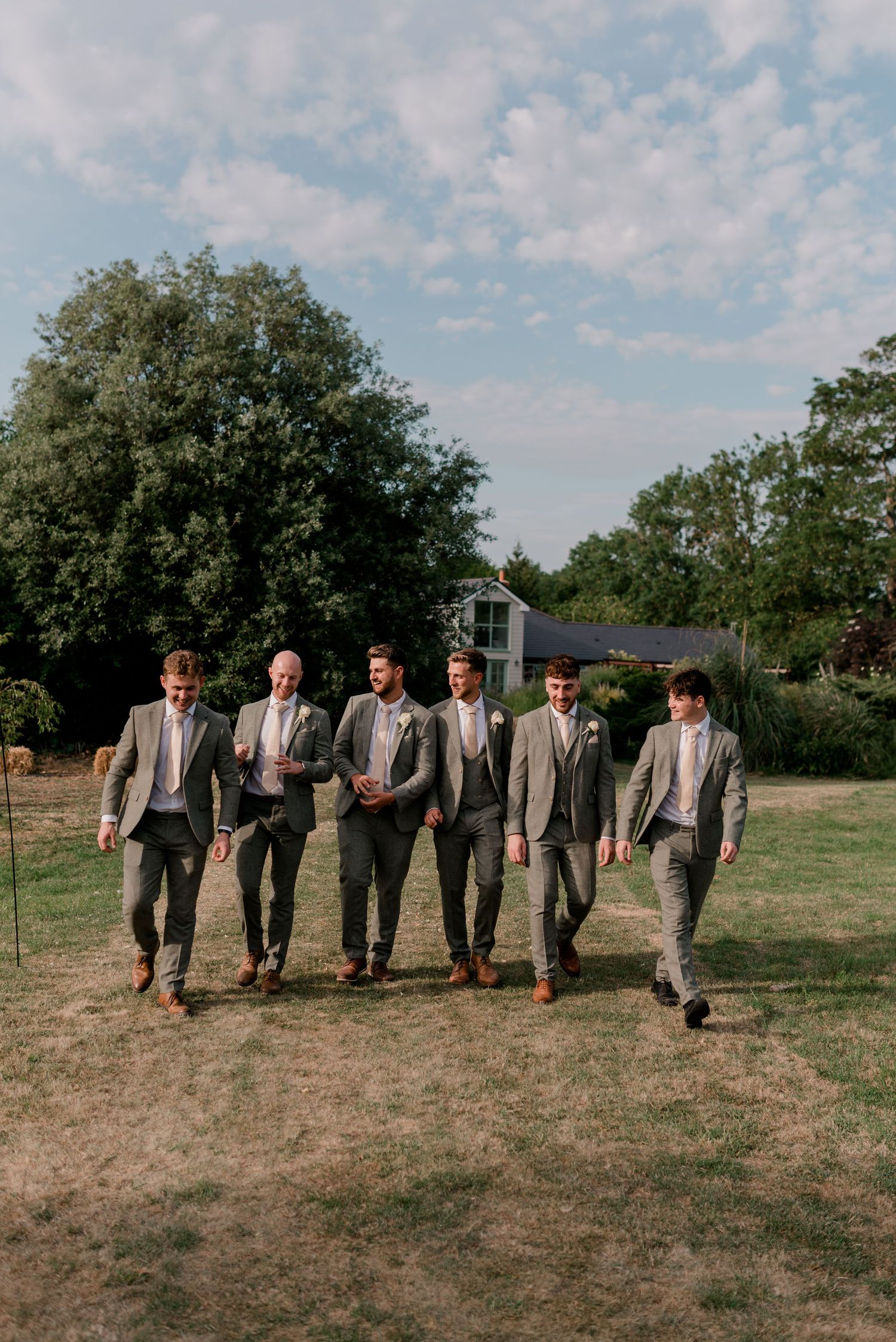 A group of groomsmen in matching gray suits walk together on a grassy field during a wedding celebration.