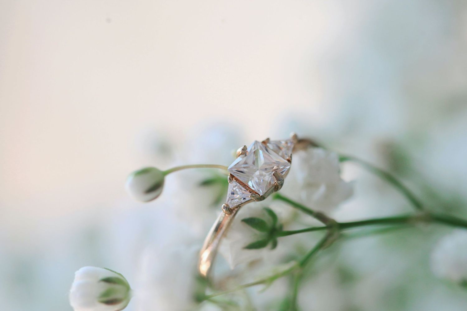 Delicate engagement ring perched on white baby's breath flowers with soft bokeh background.