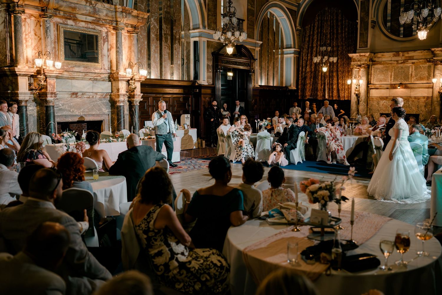 Elegant wedding reception in ornate ballroom with guests seated at round tables while bride stands in white gown.
