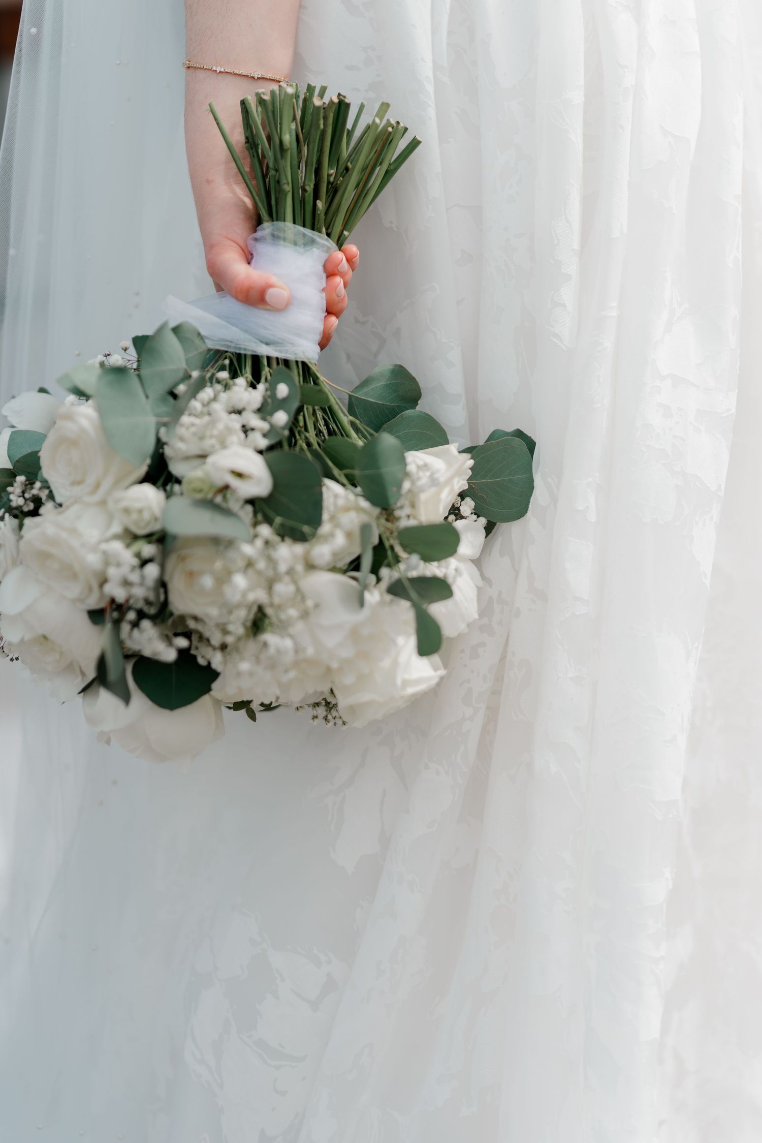 Bride holding a white wedding bouquet with roses, baby's breath, and eucalyptus against her lace gown.