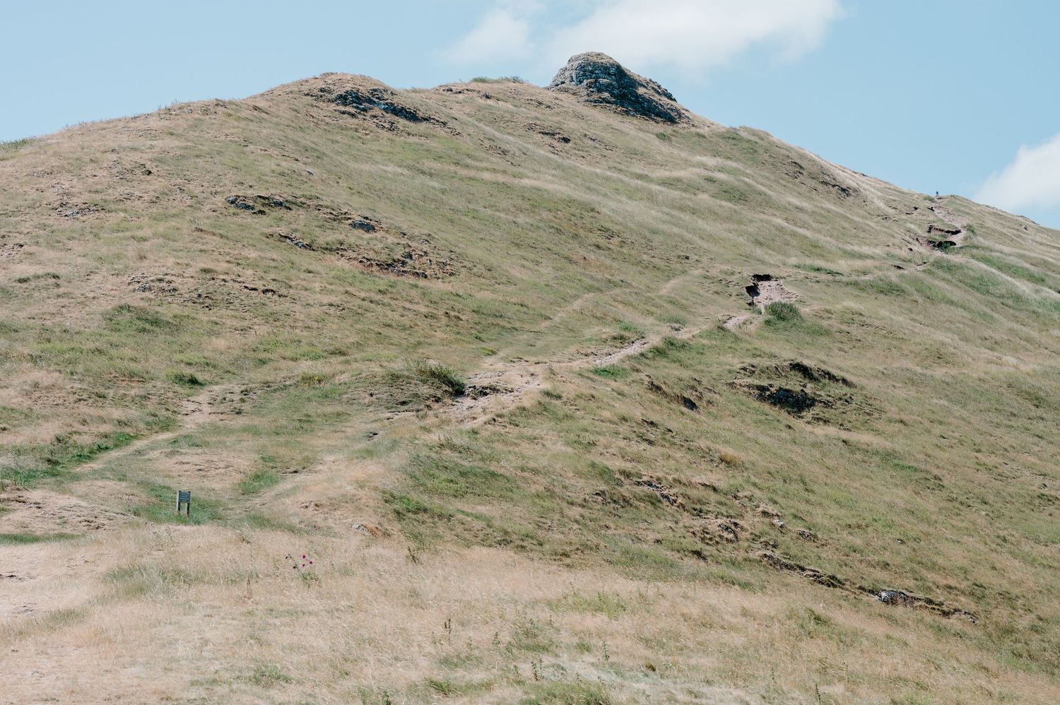 A steep grassy hillside with a rocky summit under a partly cloudy sky.