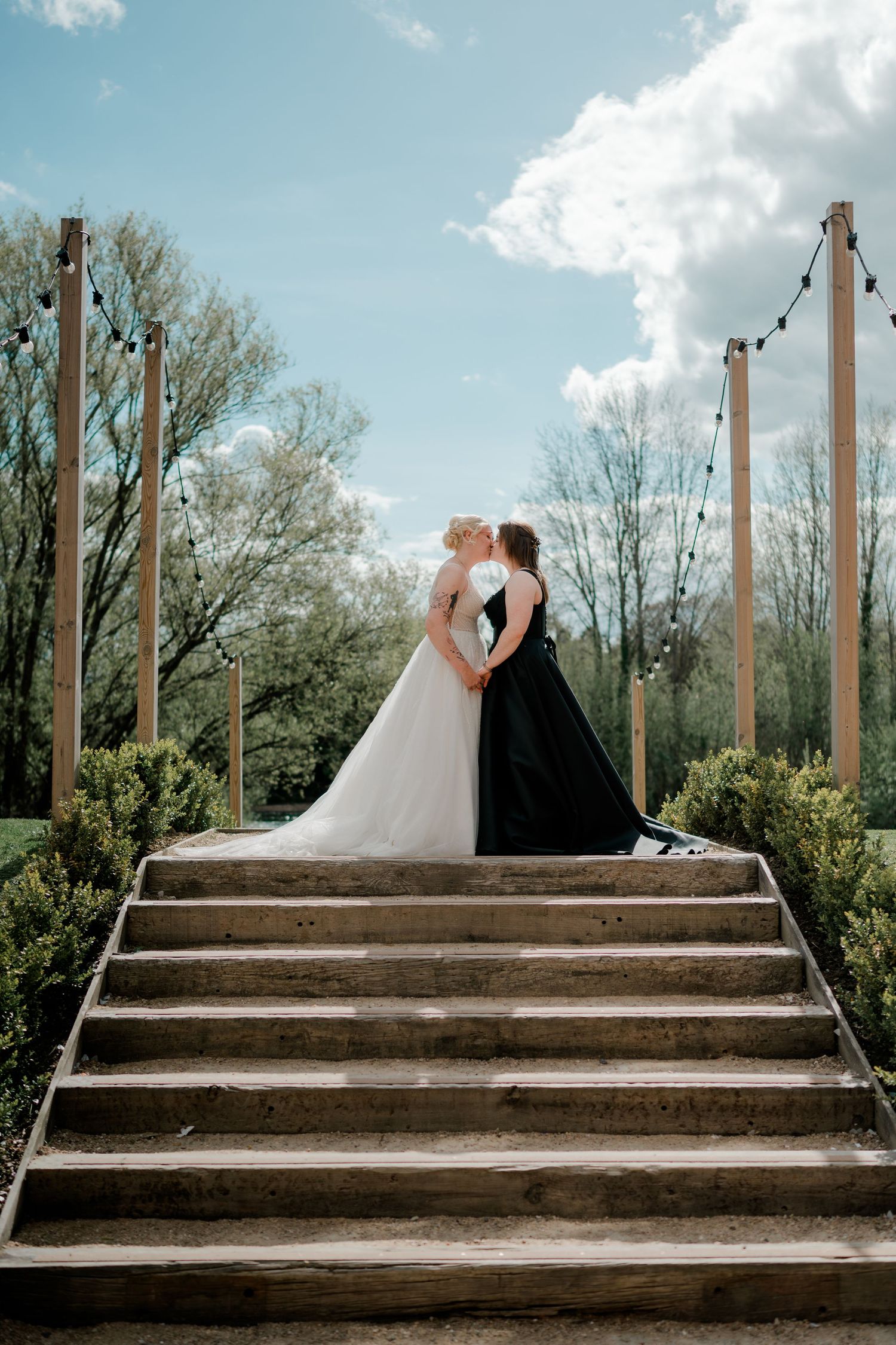 Two brides share a kiss atop wooden stairs outdoors, one in white gown and one in black dress, with string lights and trees.