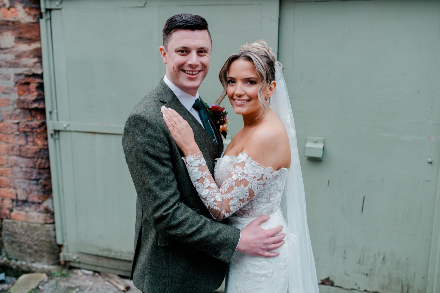 Bride in lace off-shoulder gown embraces groom in green tweed suit at wedding venue.