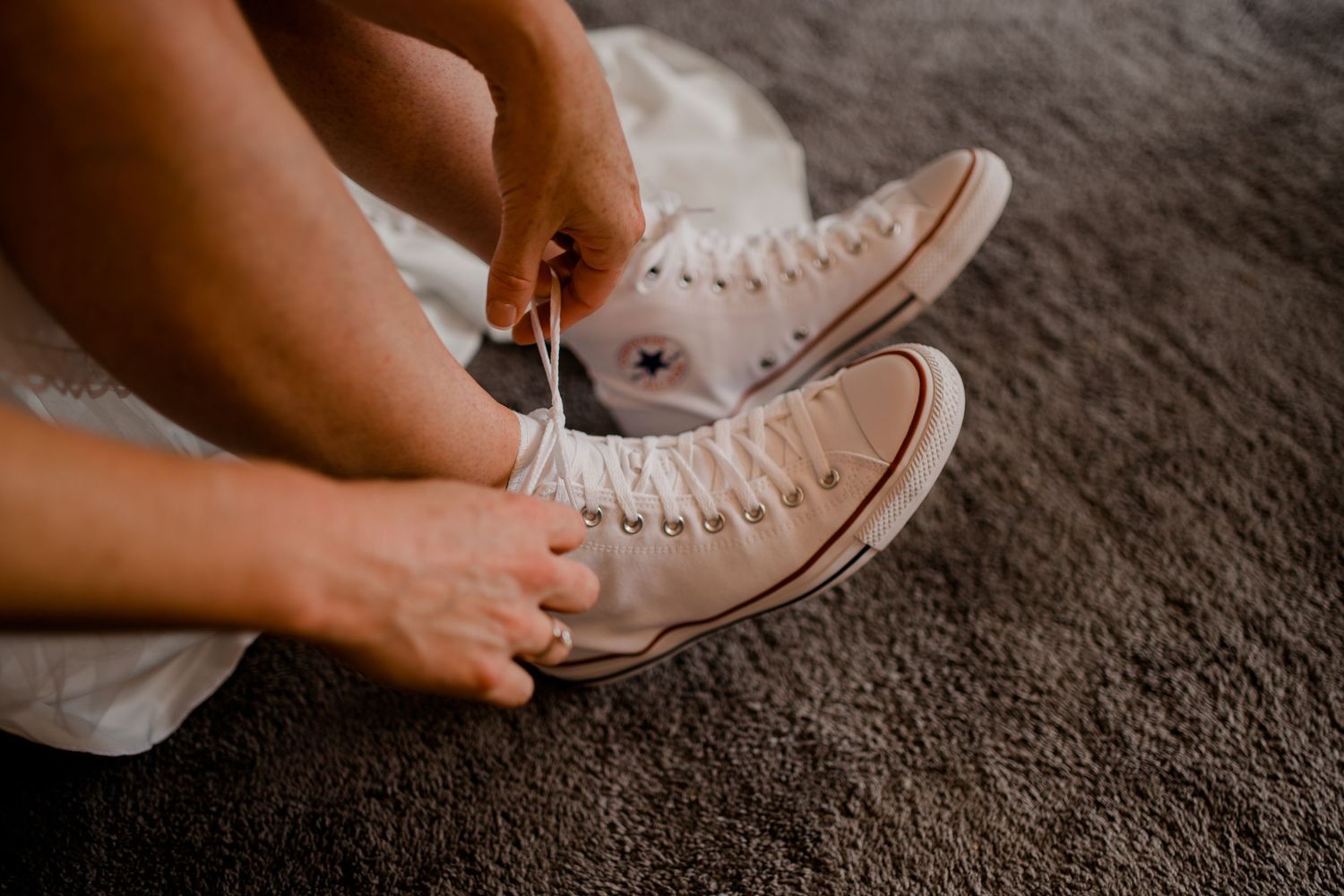 Bride tying laces on white Converse high-top sneakers while wearing a wedding dress.