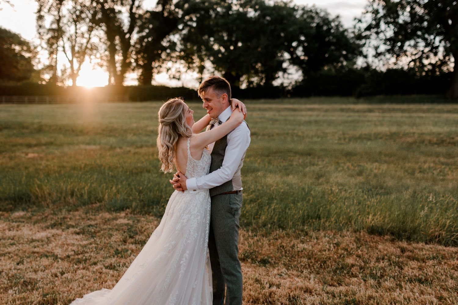 Bride and groom embrace romantically in a sunlit meadow at sunset.