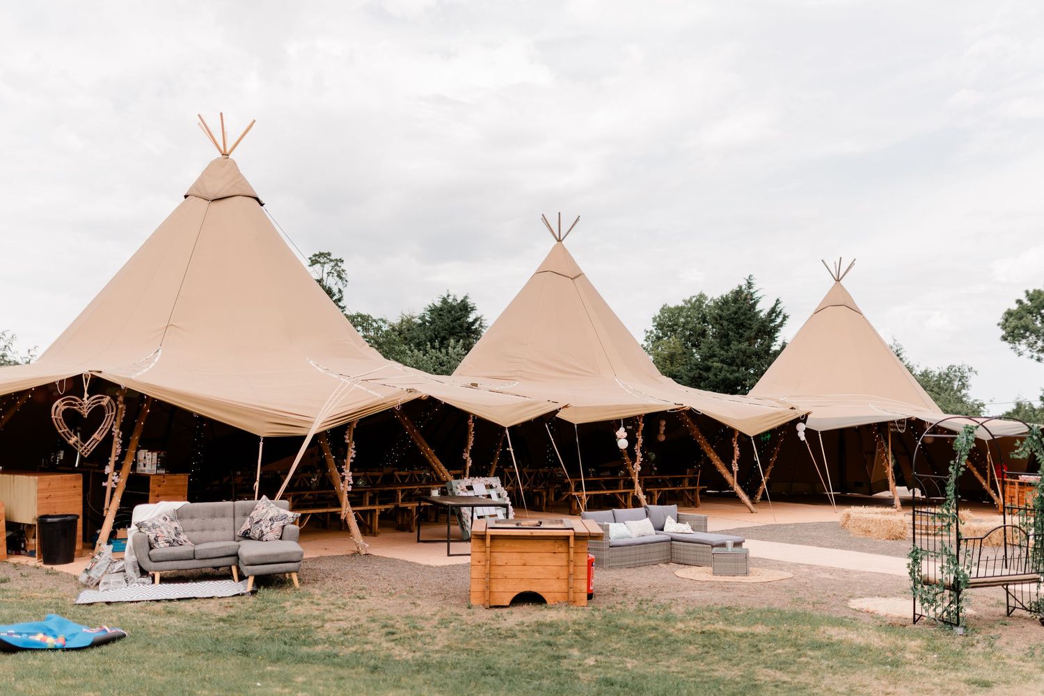 Three beige tipi-style wedding reception tents with lounge furniture set up on grass under cloudy skies.