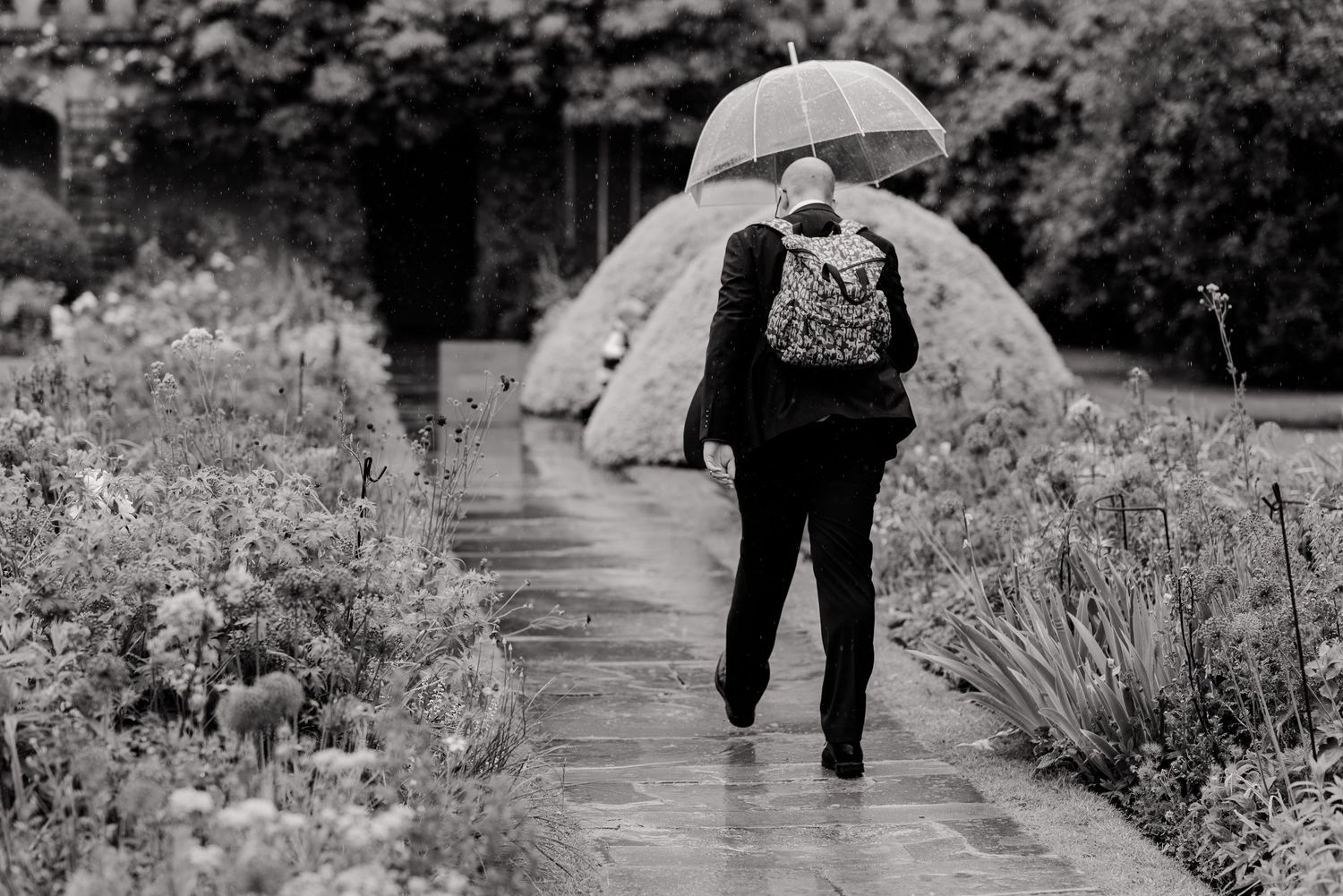 Person walking on garden path carrying umbrella in black and white photograph.
