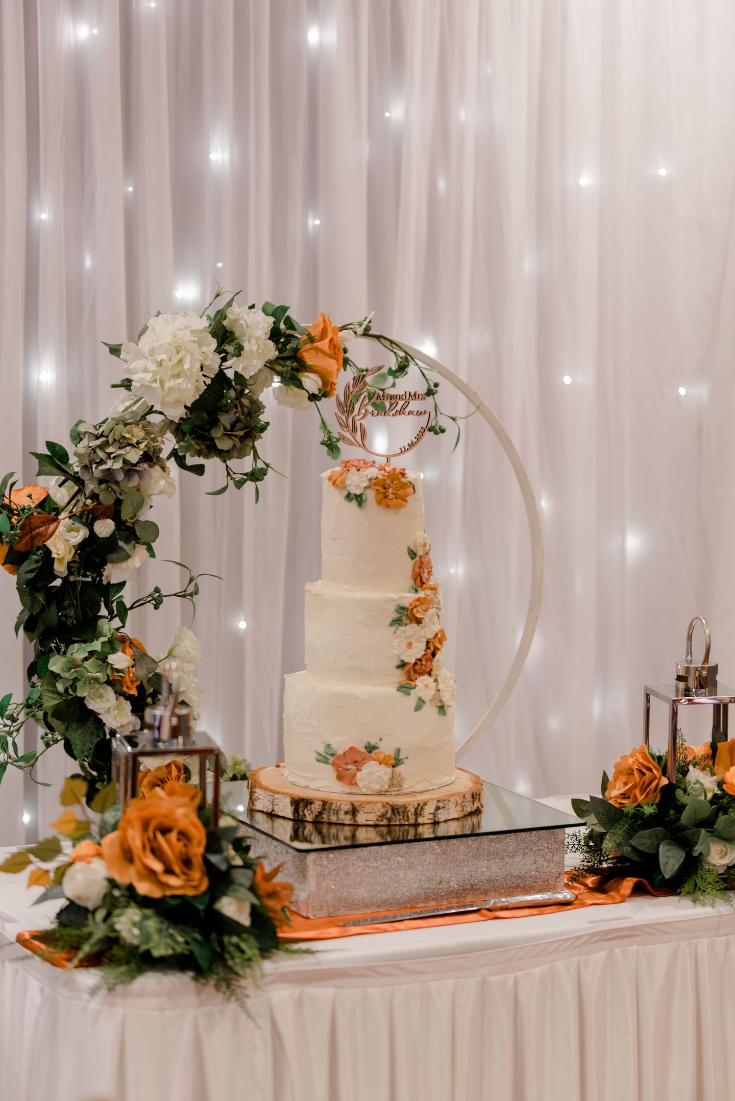 Elegant wedding cake display with circular floral arch and orange roses against shimmering white backdrop with fairy lights.