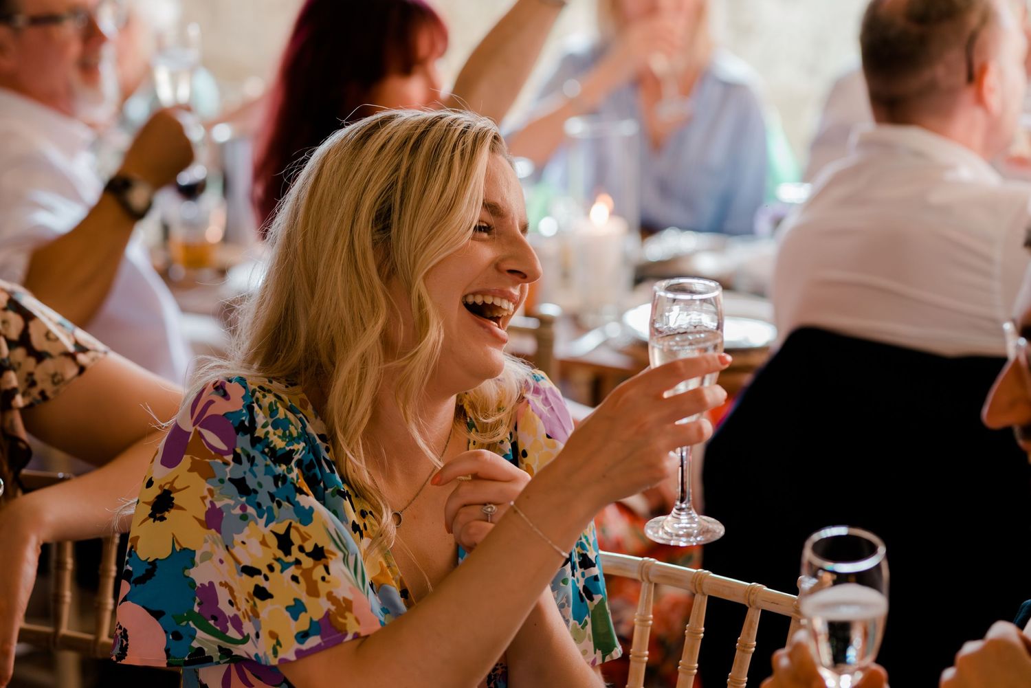 Wedding reception guests laughing and enjoying drinks at tables with exposed brick wall backdrop.