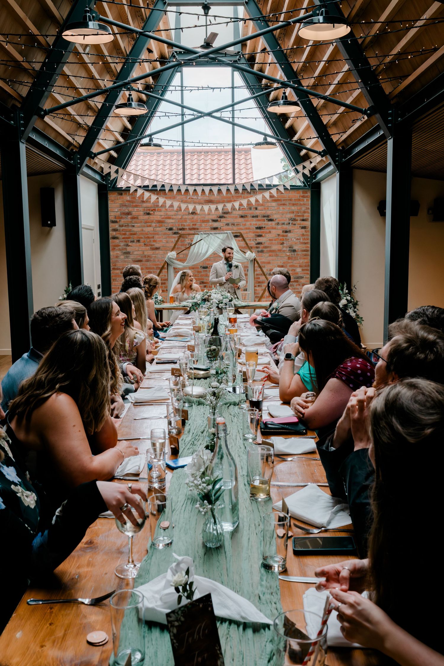 Long wedding reception dinner table with greenery runner, guests seated on both sides in an industrial brick venue.