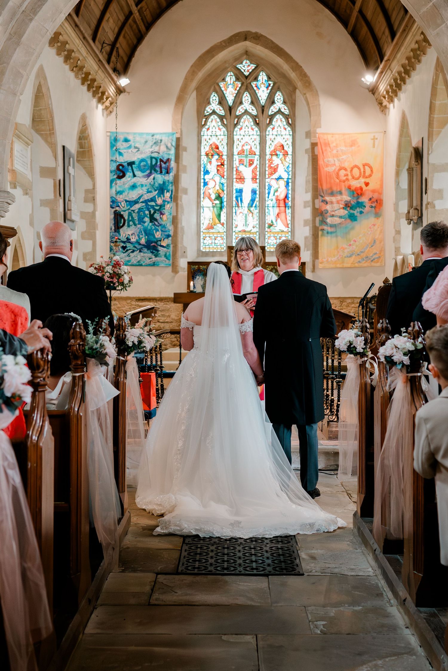 Bride and groom stand at church altar during wedding ceremony with stained glass windows and colorful banners behind officiant.