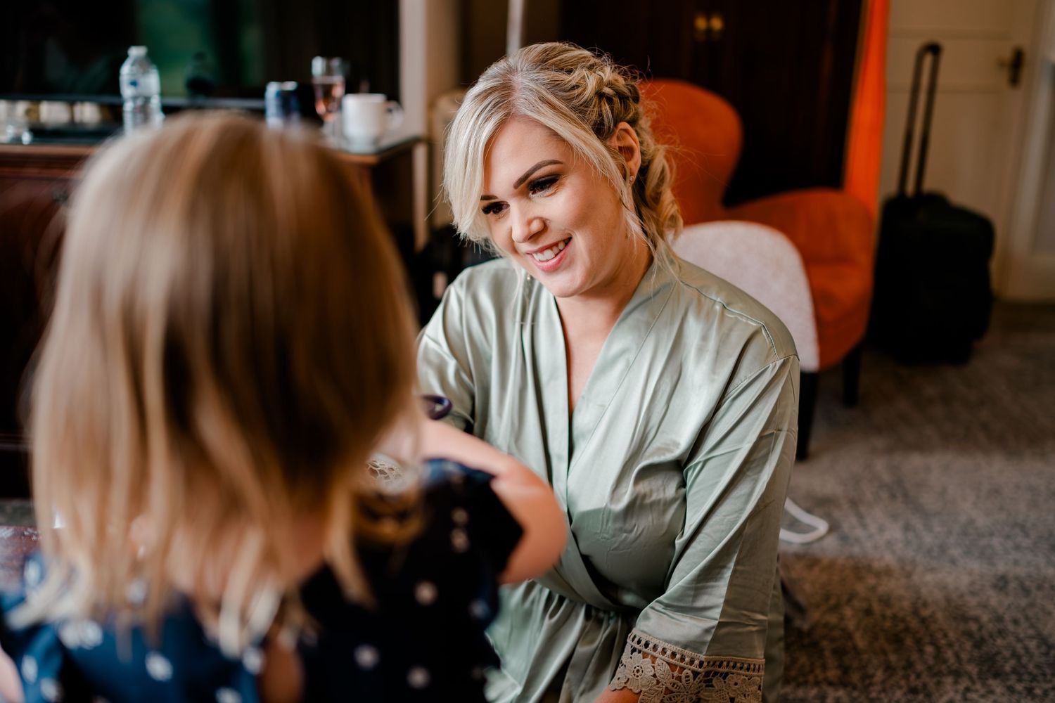 Smiling woman with an elegant updo wearing a green satin robe kneels to chat with a little girl in a polka dot dress in a cozy room.