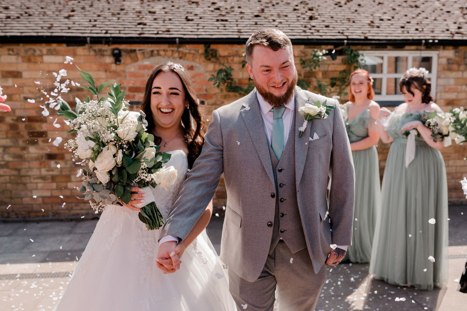 Wedding party members in gray suits and sage green dresses standing against brick wall.