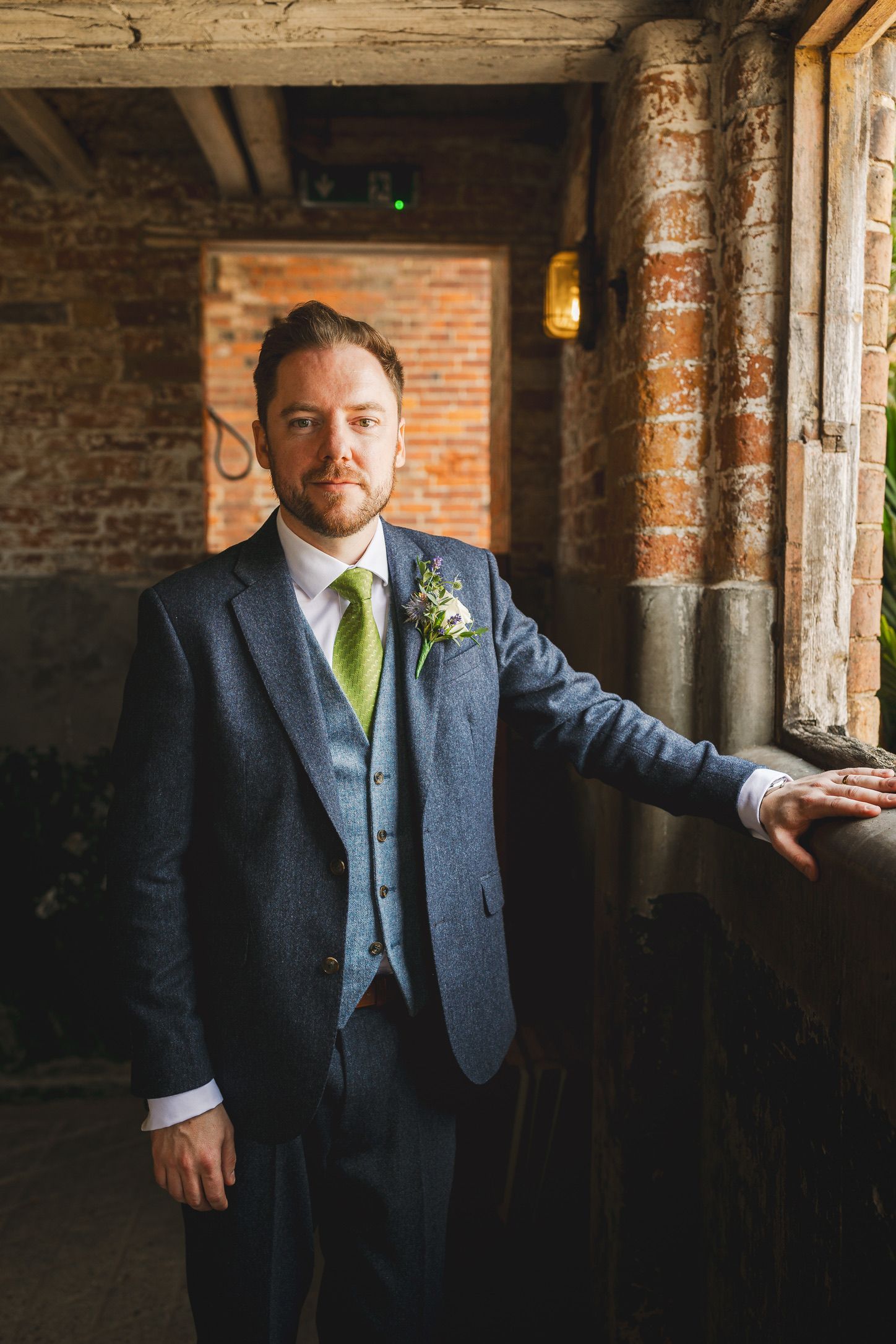 Dapper groom in grey tweed suit with green tie posing in rustic brick venue.