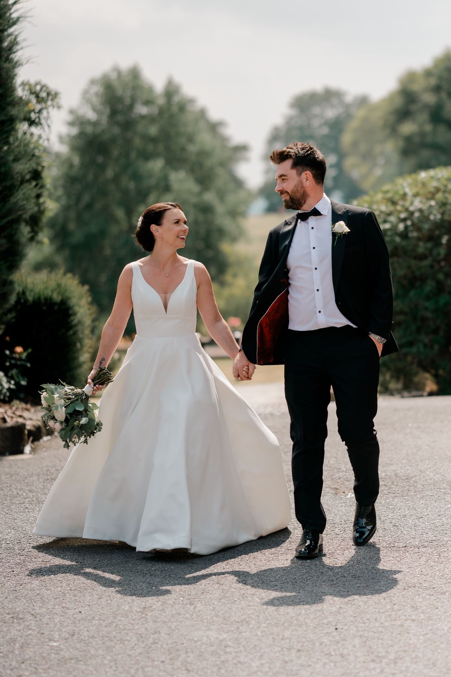 Bride and groom walking hand in hand outdoors, smiling at each other, wearing wedding dress and black tuxedo.