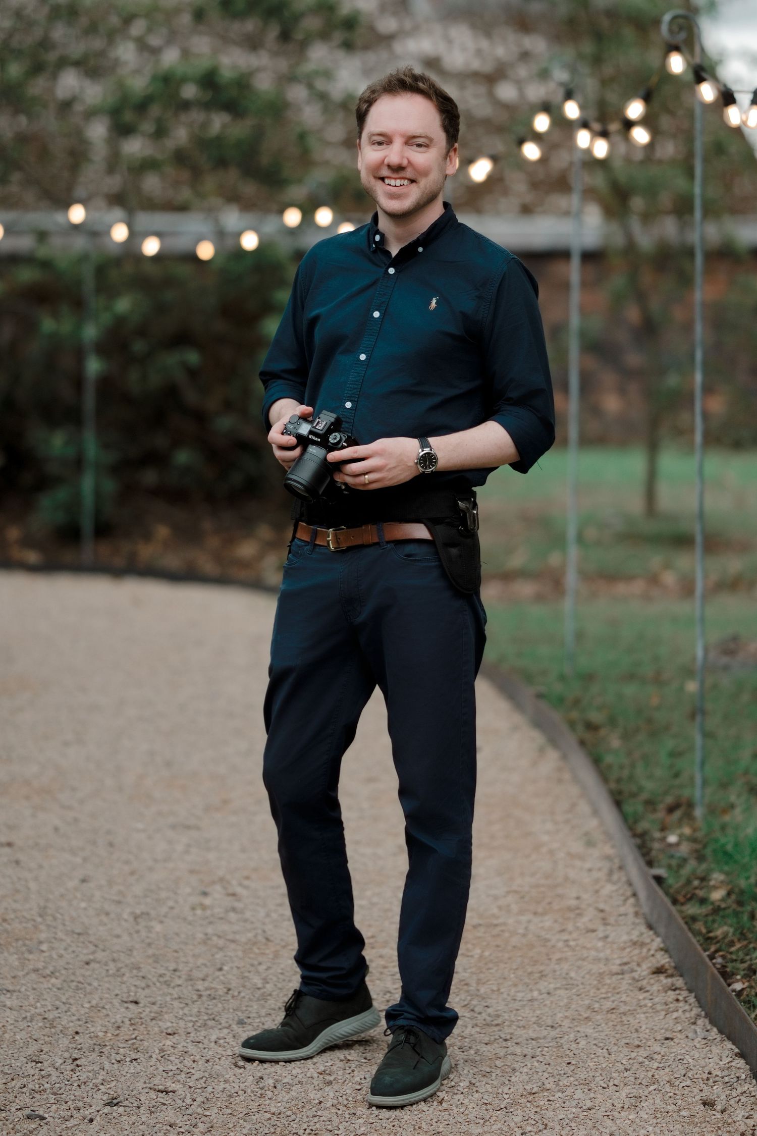 Smiling man in dark teal shirt holding a camera on a gravel path with string lights in background.