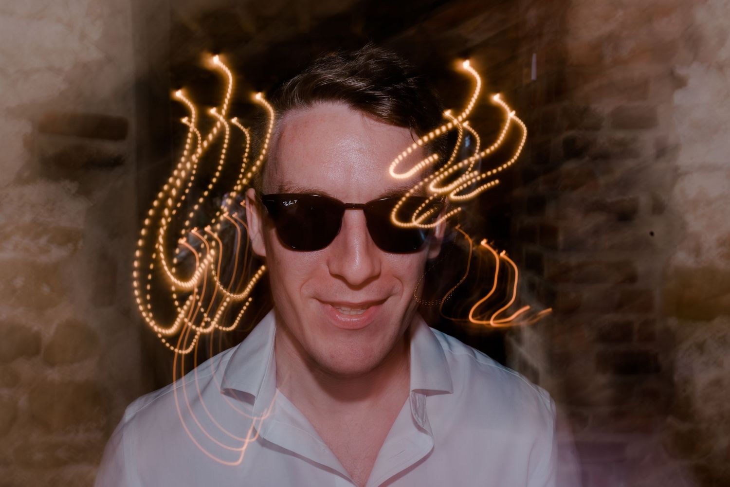 Party guests posing with sunglasses and light trails in front of brick wall at nighttime event.
