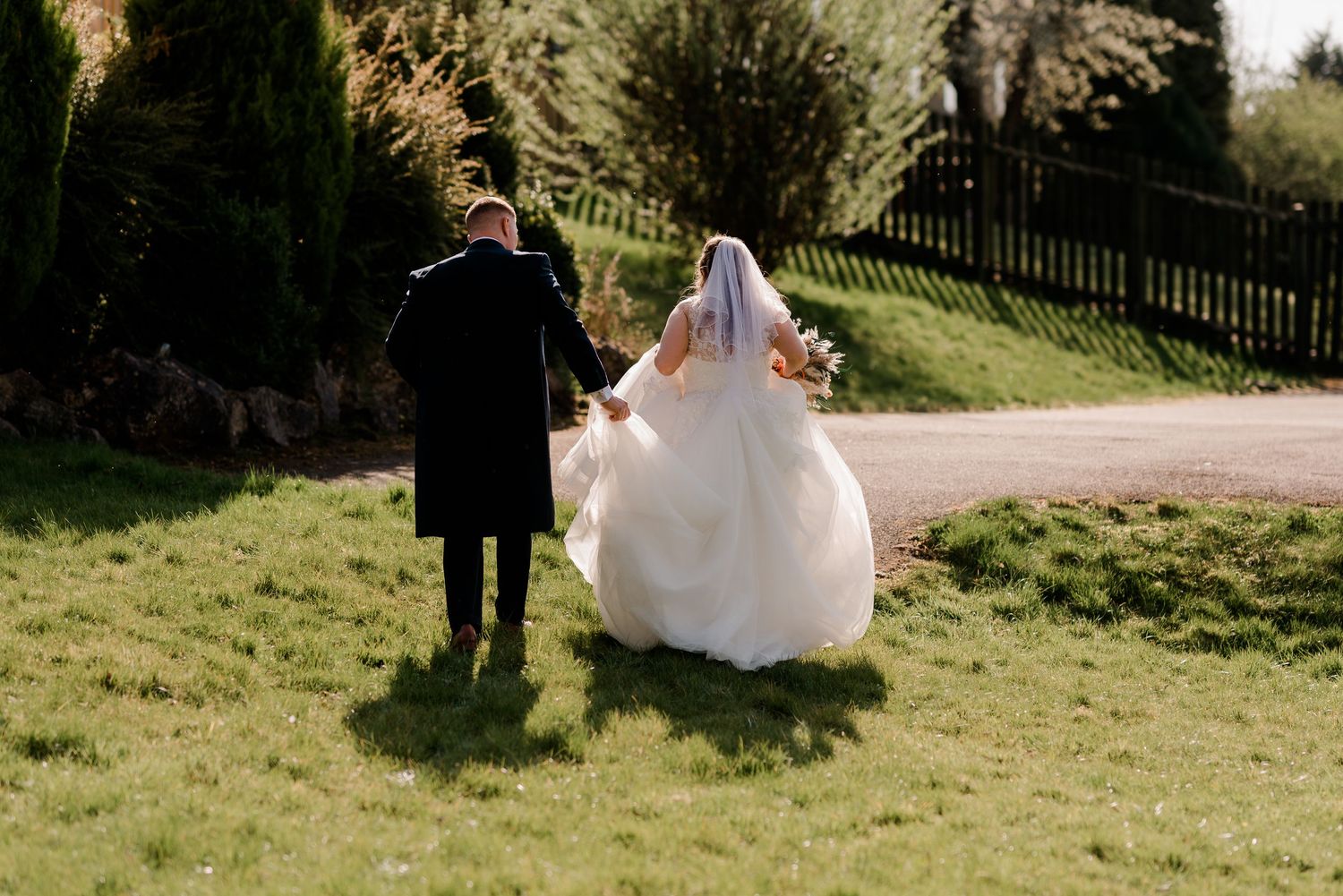 Wedding couple walking down a garden path on a sunny day with the bride wearing a flowing white gown.