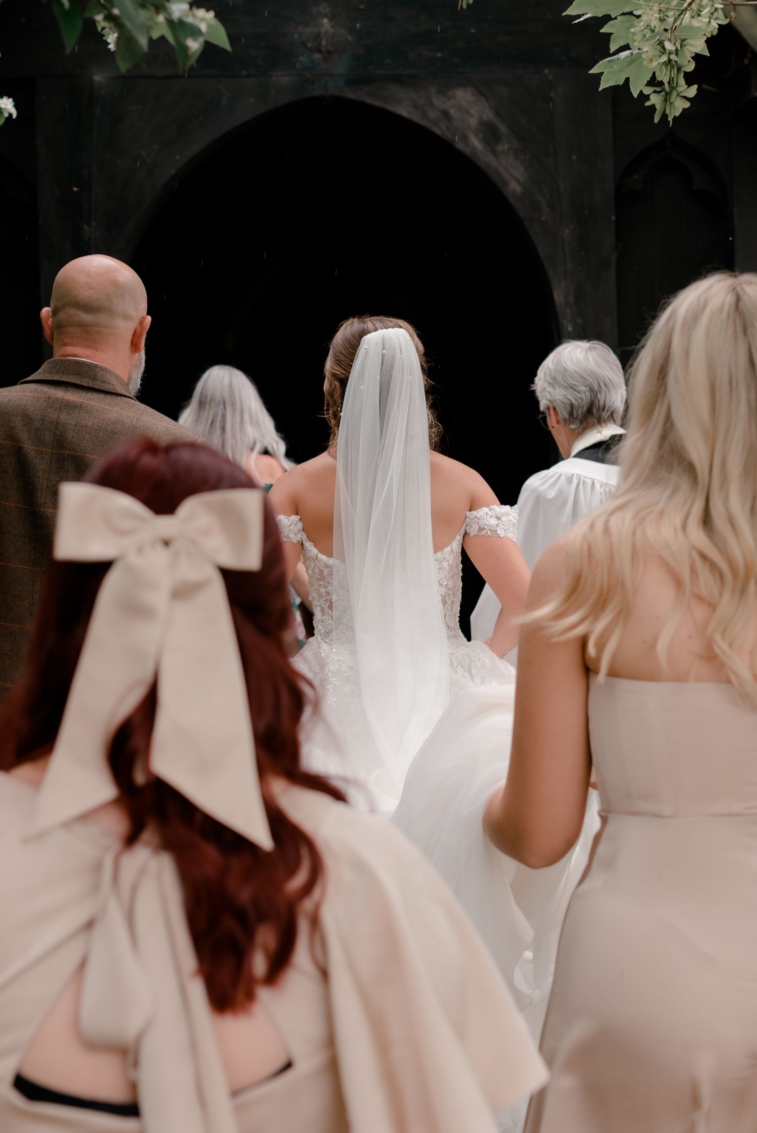 Bride and wedding party in white and blush dresses enter through an archway with a dramatic veil trailing behind.