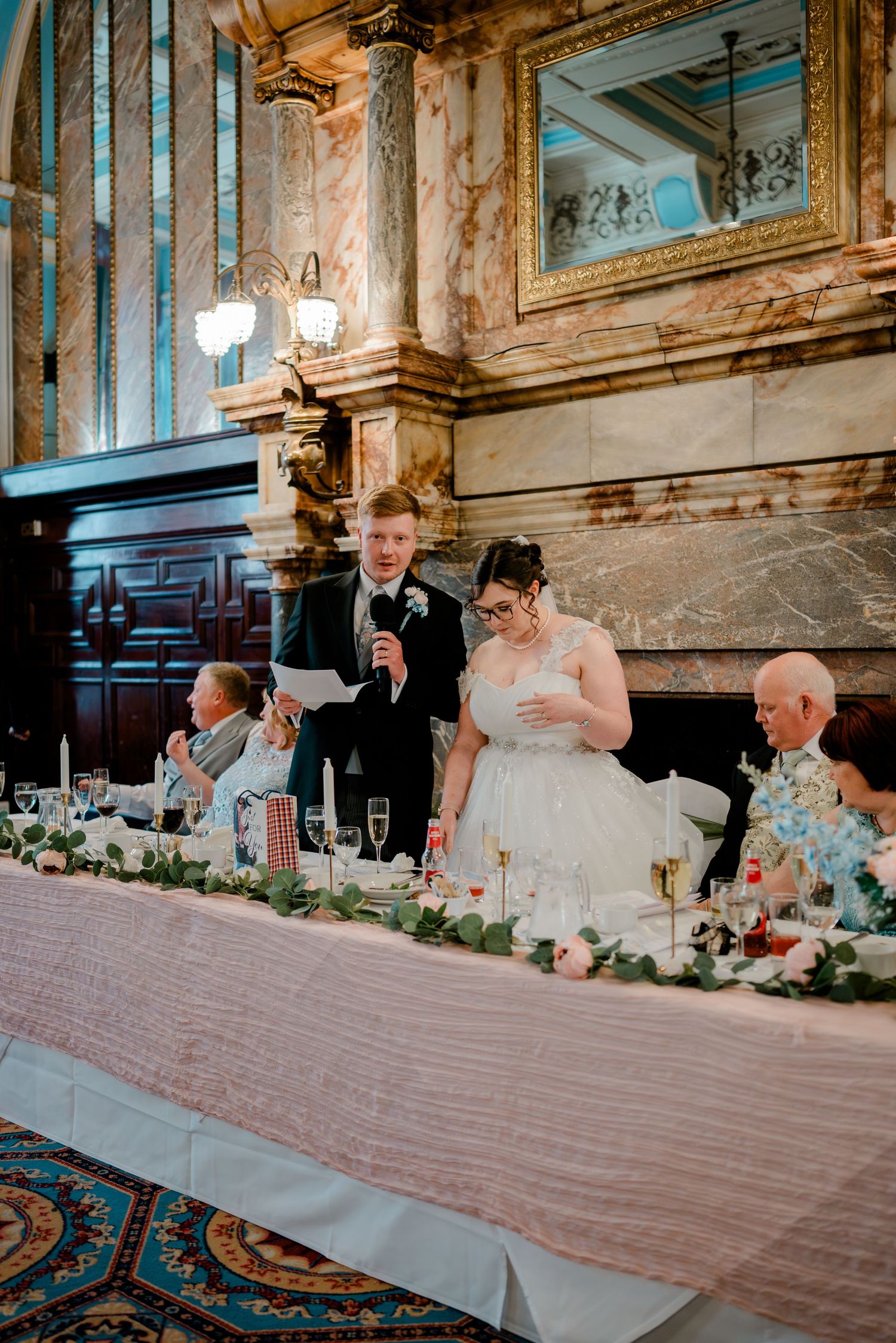 Bride and groom giving speech at elegant wedding reception with marble fireplace and floral table garland.