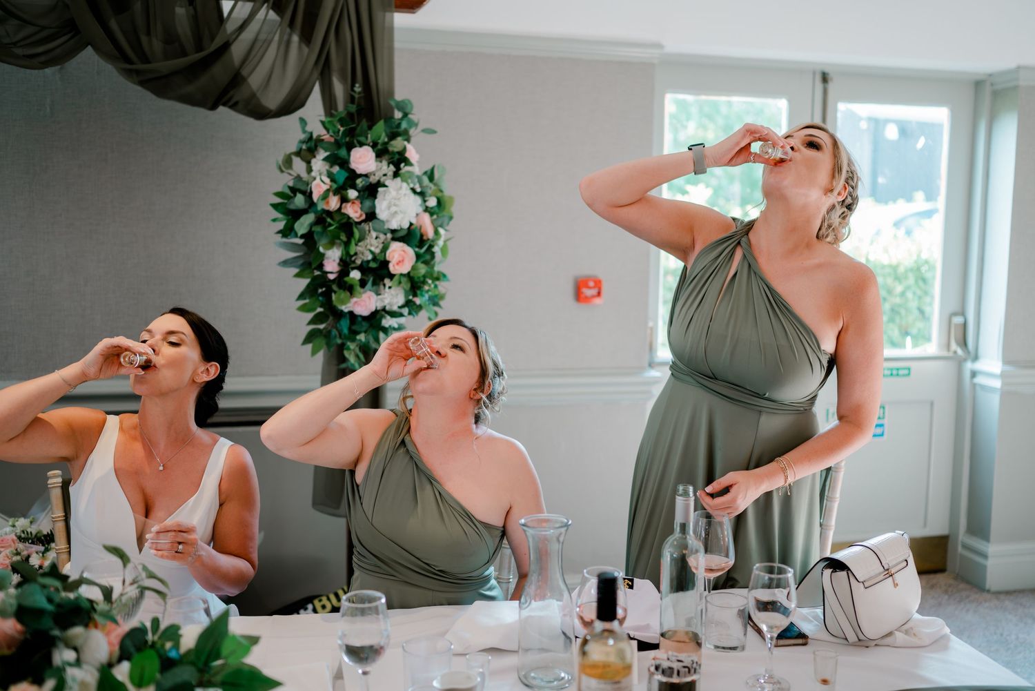 Three bridesmaids in olive green dresses take shots at a wedding reception table beside a floral wreath and glassware.