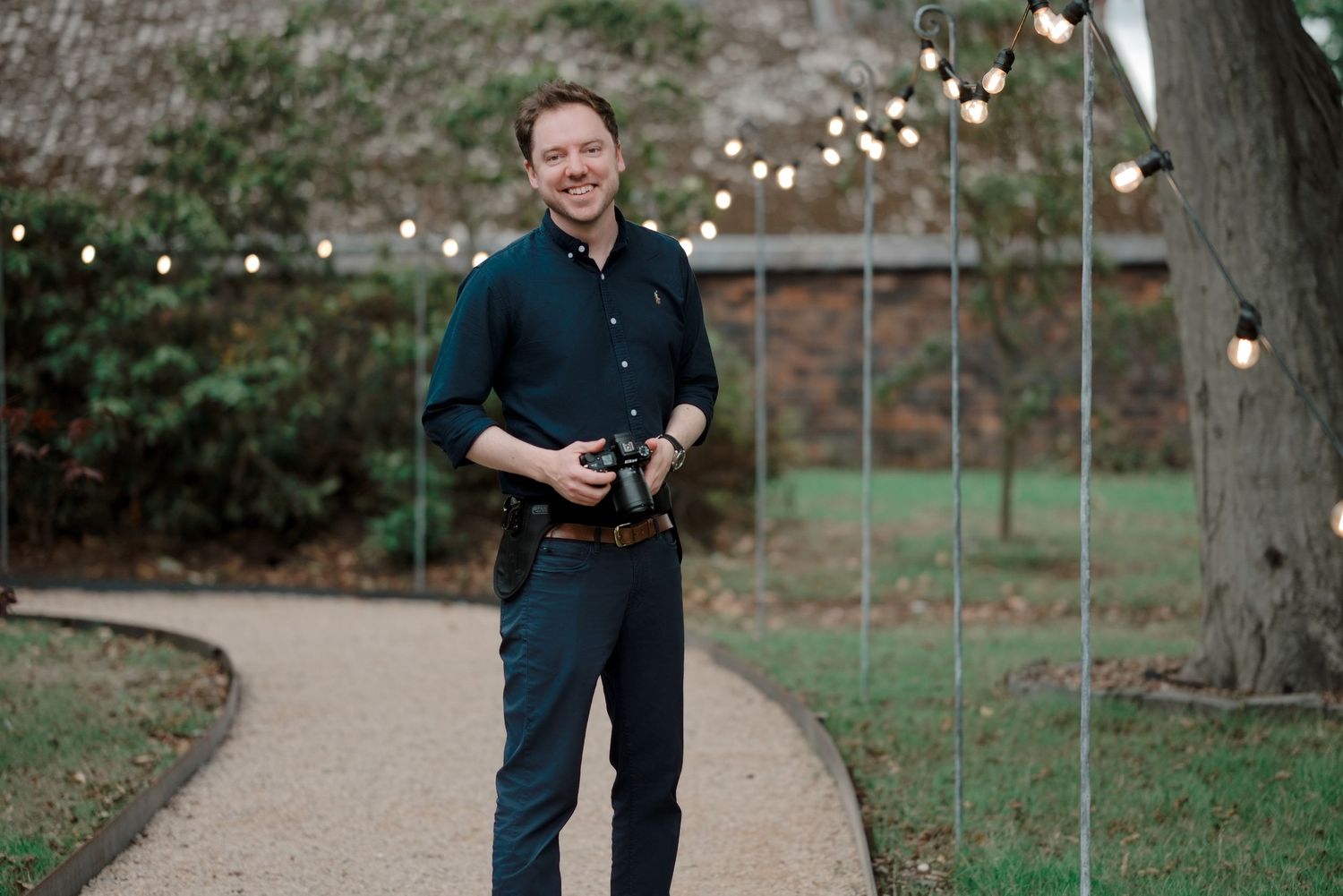 Smiling man holding a camera on a garden path with string lights, wearing a dark shirt and navy pants.