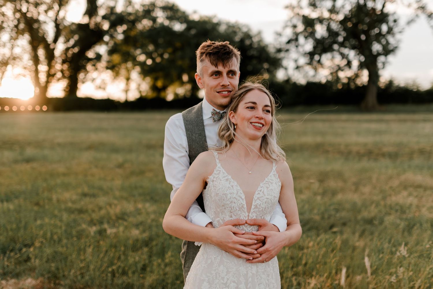 Groom embraces bride in lace V-neck gown during golden sunset in a scenic outdoor field setting.