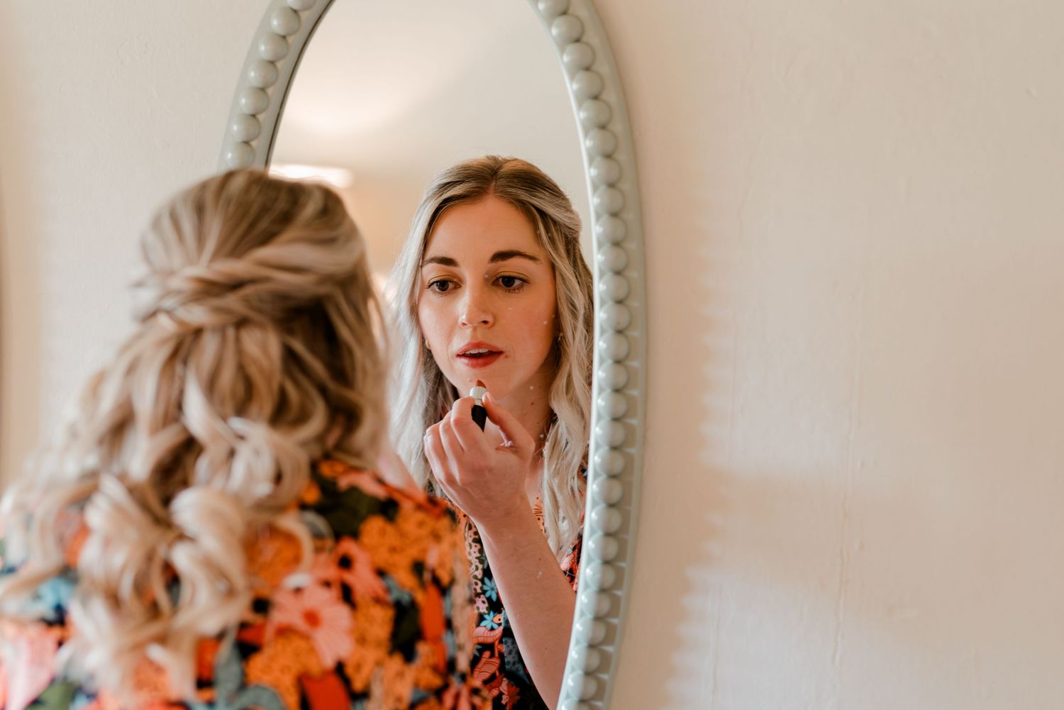 Reflection in an ornate oval mirror with floral orange dress and delicate makeup application.