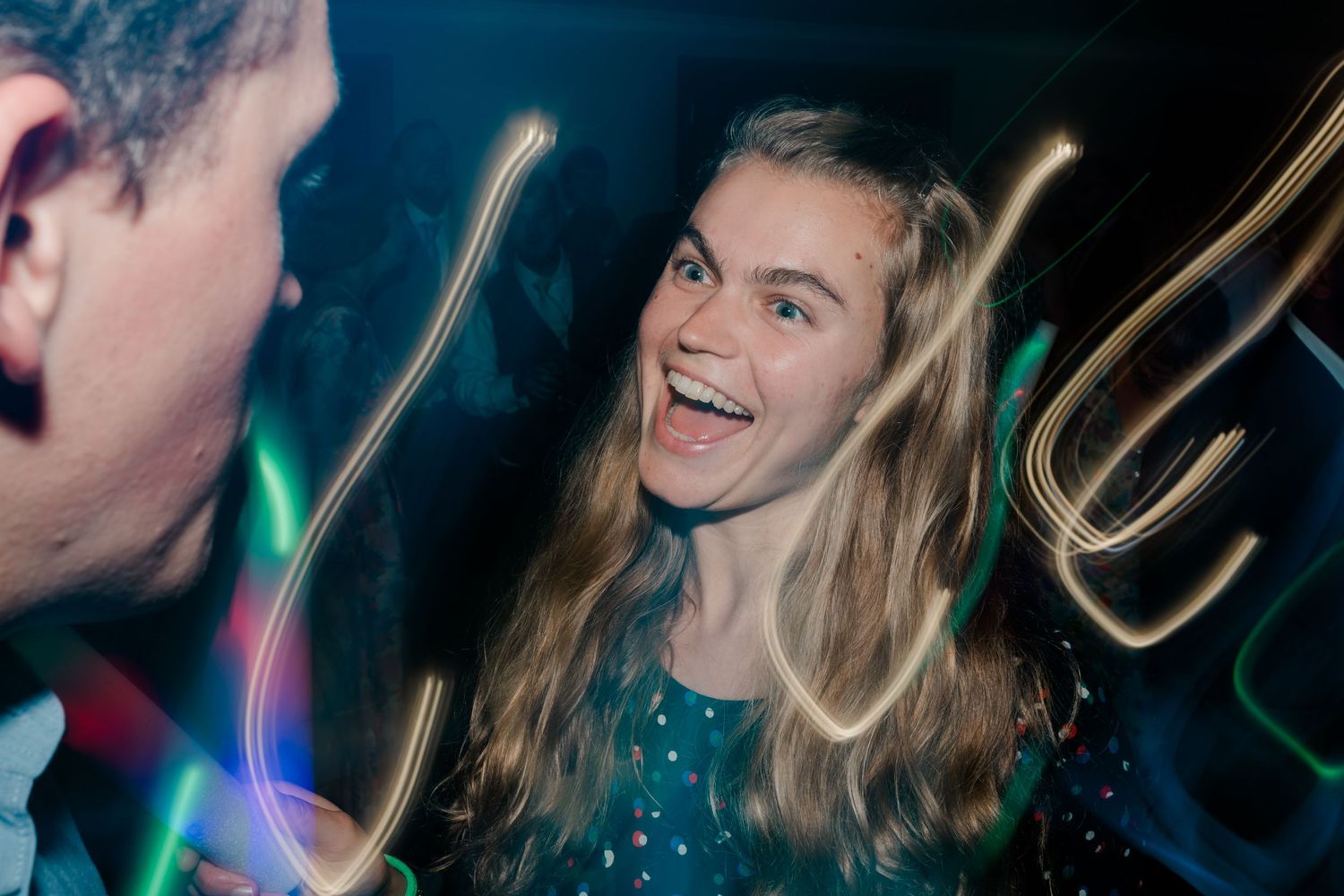 Wedding guests dance and celebrate on a dimly lit dance floor with colorful lights creating a festive atmosphere.