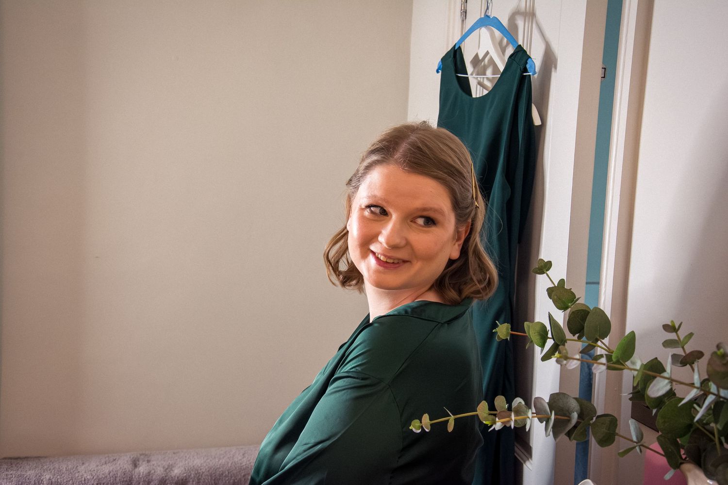 Someone wearing a dark green dress poses in a bright interior room with hanging clothes.