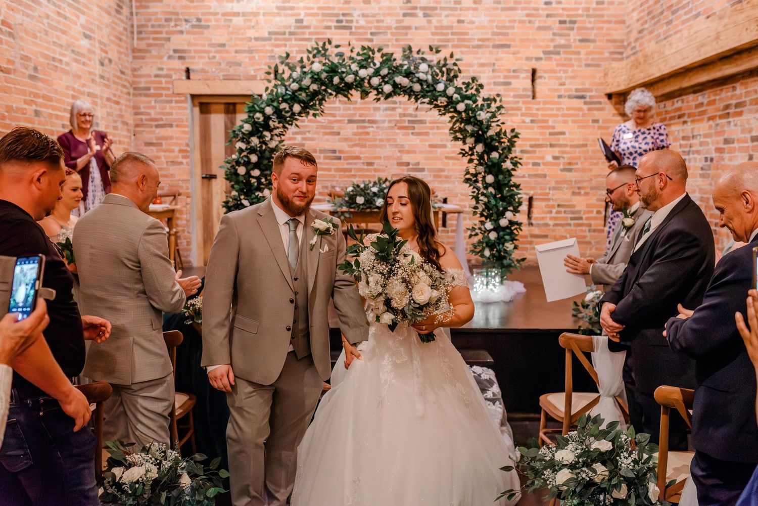 Wedding celebration moment under floral arch with exposed brick backdrop.