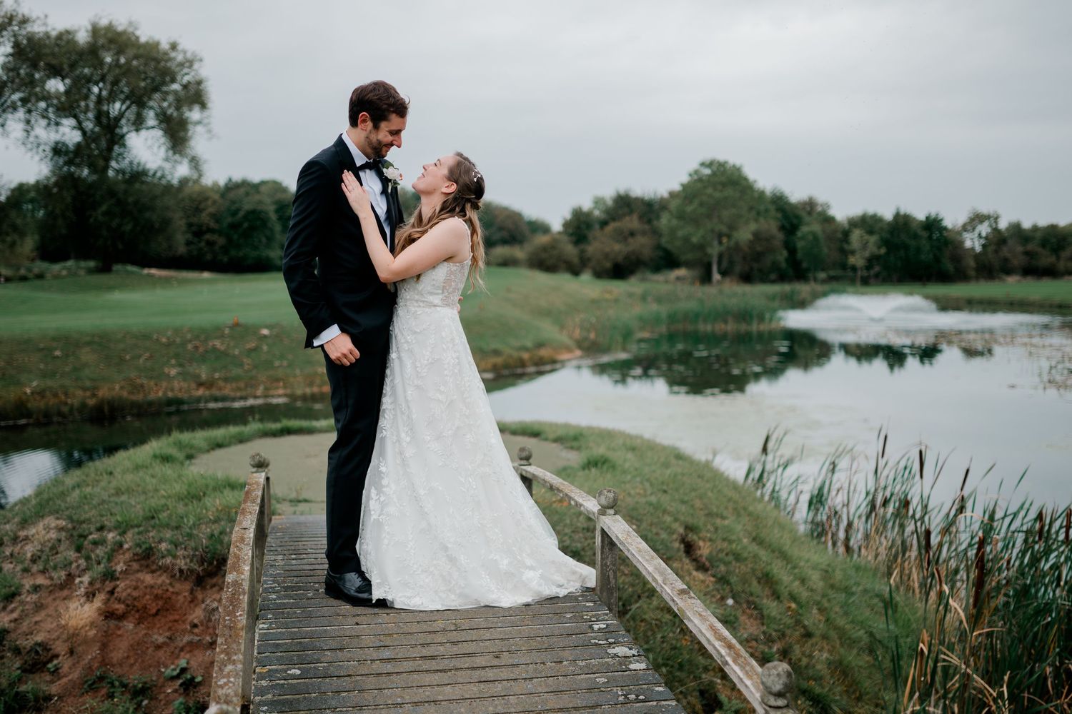 Romantic wedding couple embraces on a golf course overlooking a serene pond on an overcast day.