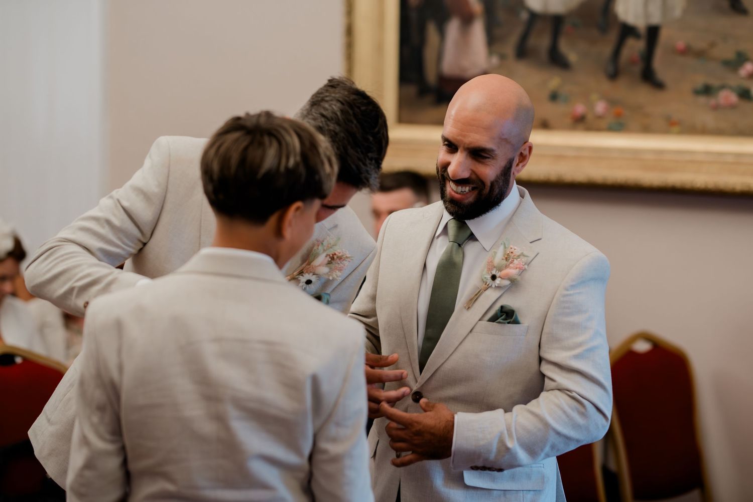 Groomsmen in beige suits with green ties and boutonnieres laughing together before a wedding ceremony.