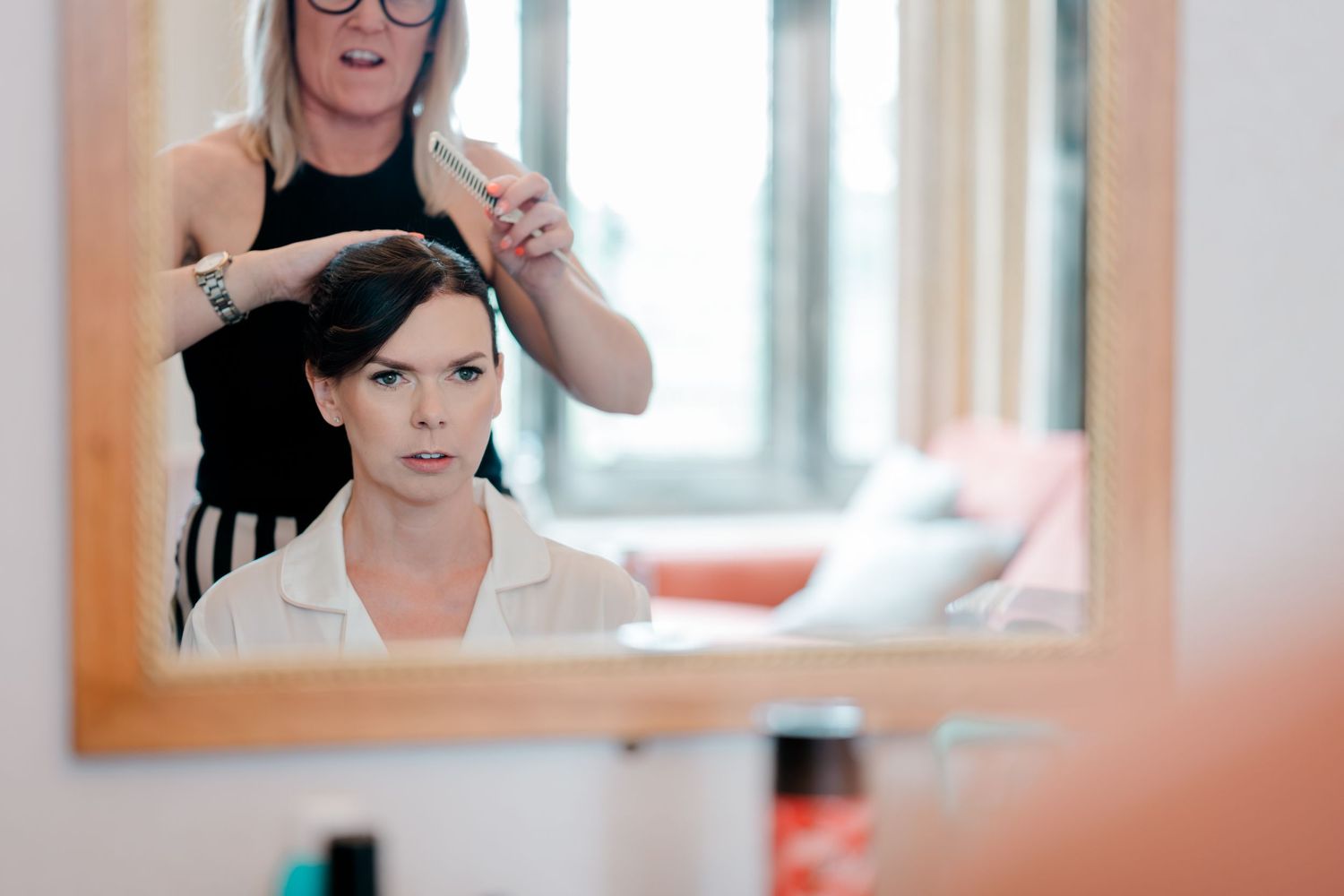 A bride sits before a wooden-framed mirror while a hairstylist styles her dark hair during wedding morning preparations.