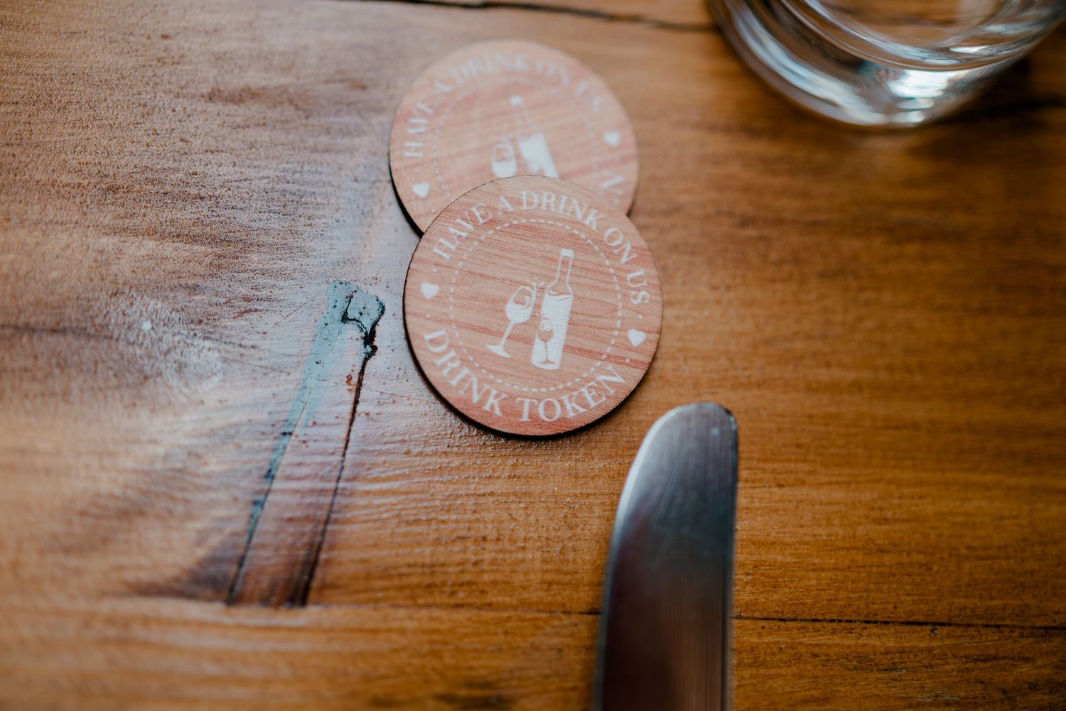 Two round pink wooden coasters with white text on a wooden table beside a knife and glass.