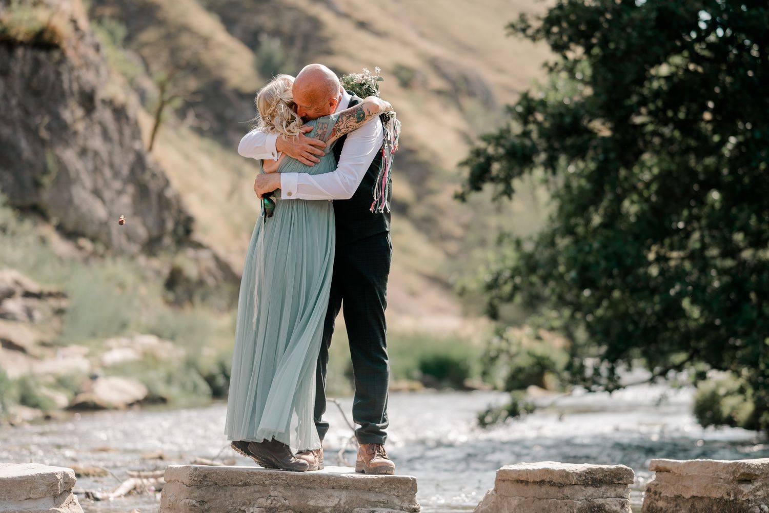 Couple embracing in a romantic outdoor wedding ceremony near a river with scenic mountain backdrop.