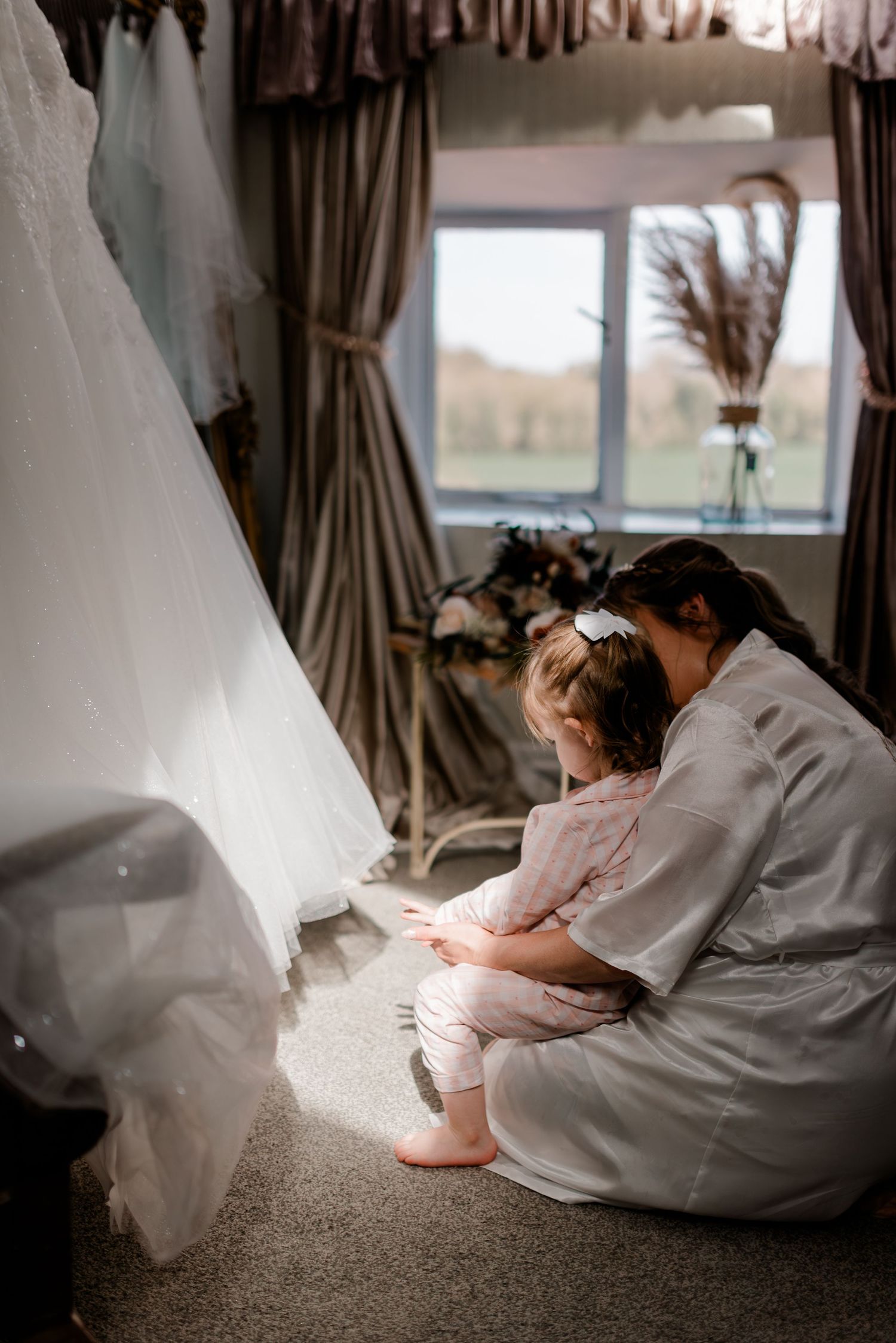 A tender moment of getting ready on a wedding day, with a white dress visible in the foreground and soft sunlight through a window.