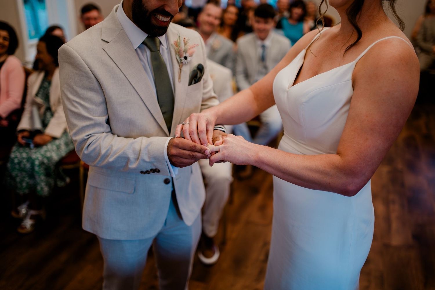 A couple exchanges wedding rings during an intimate ceremony with guests looking on in the background.