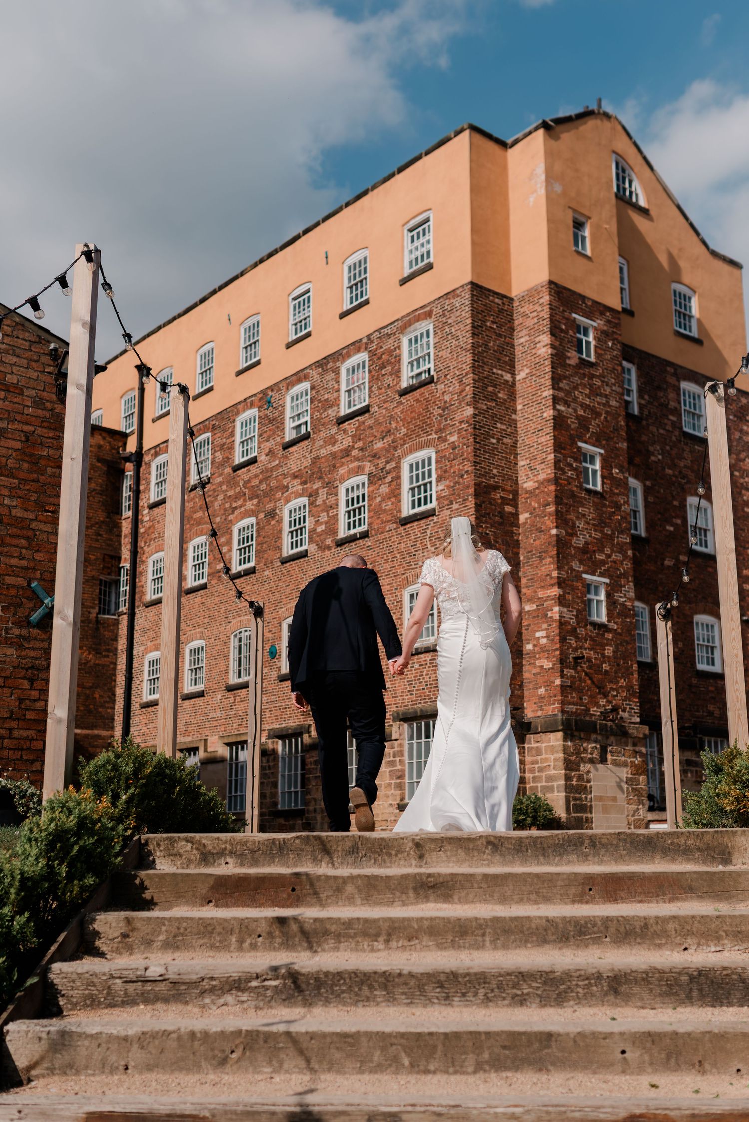 Bride and groom holding hands walking up stone steps toward a large red brick wedding venue building.