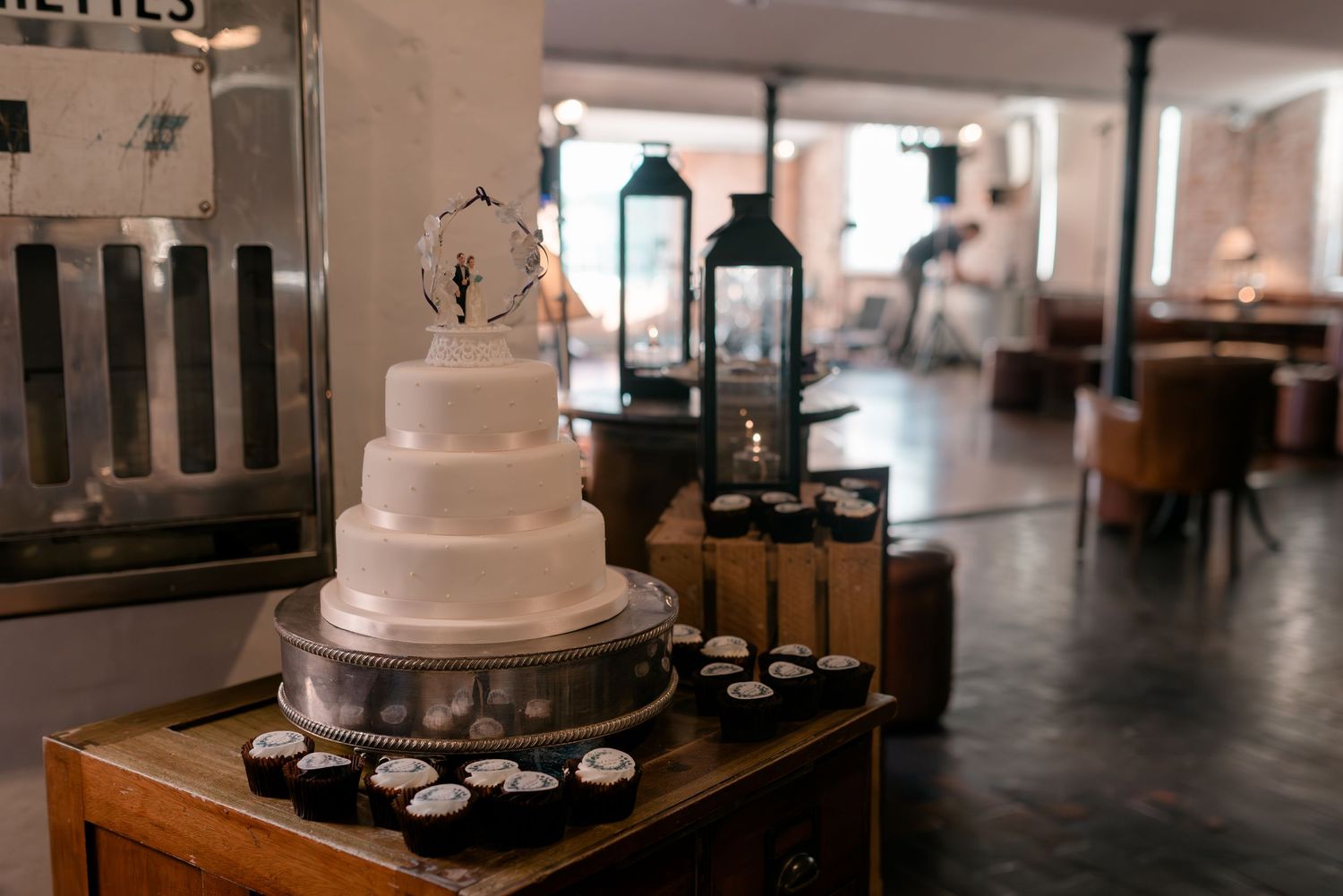 Elegant four-tier white wedding cake displayed on wooden table with cupcakes and lanterns in rustic venue.